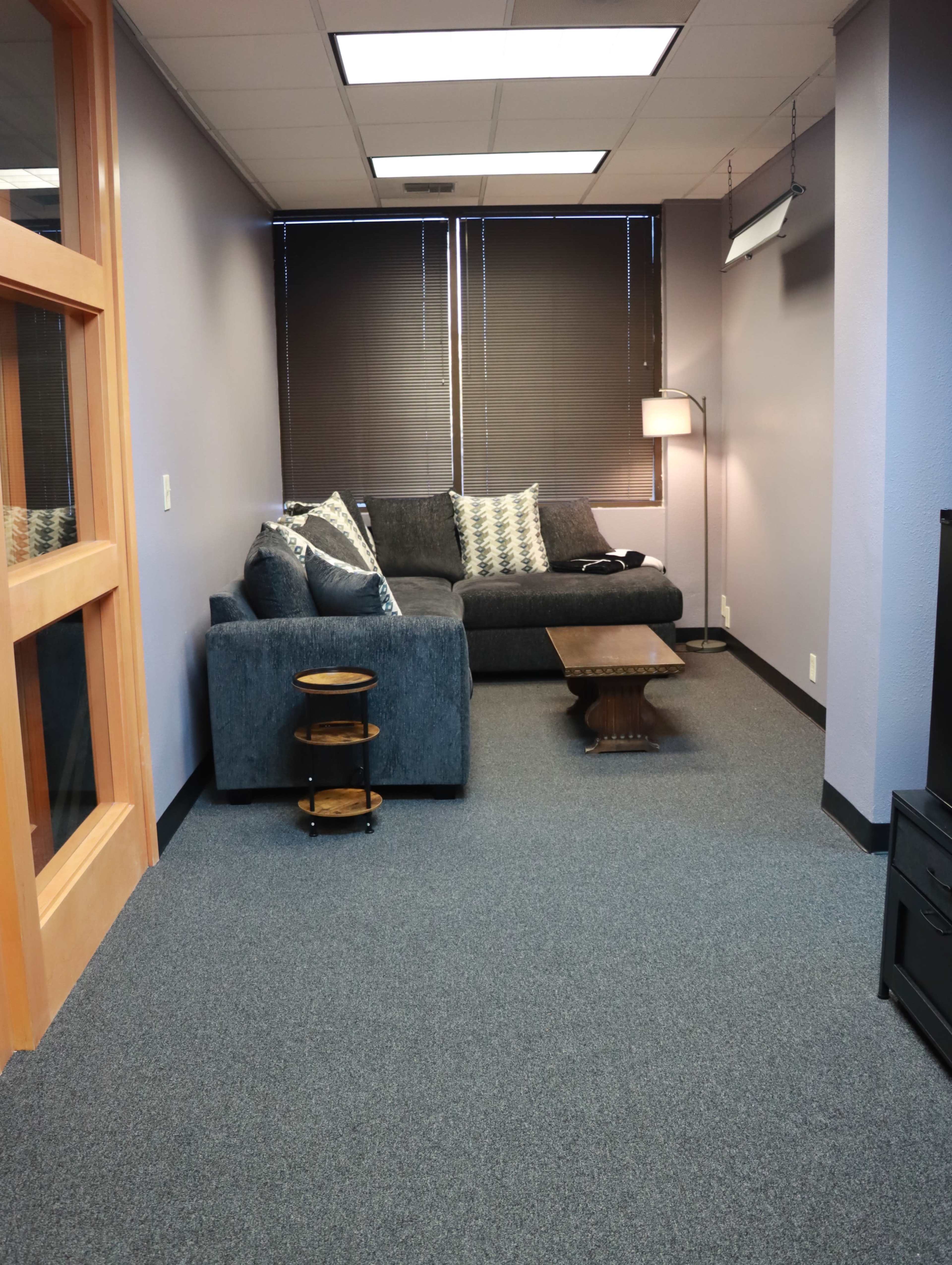 The image shows a small office lounge area with a gray couch, a wooden coffee table, and a light from a floor lamp, all situated against a wall with dark blinds.