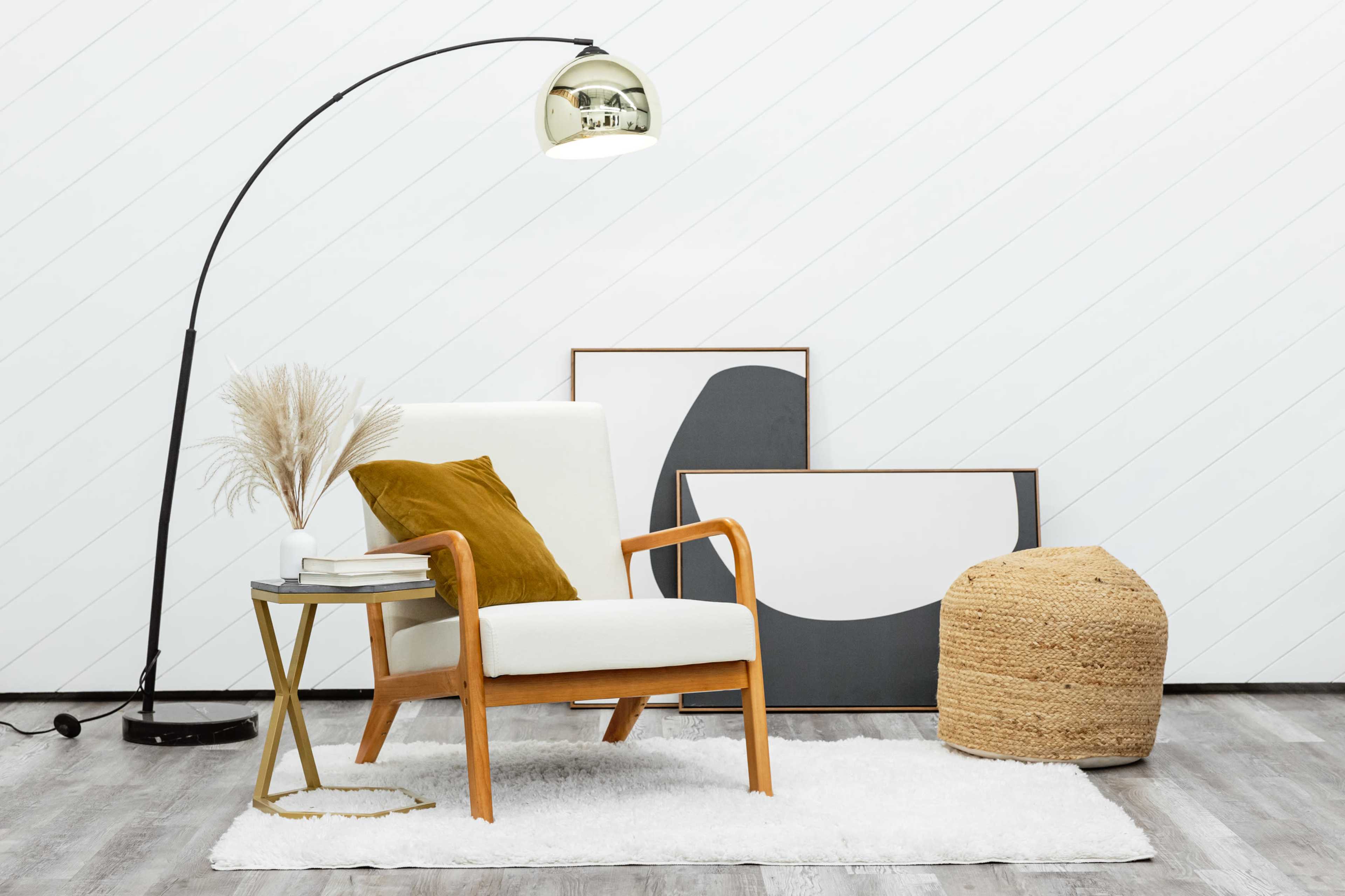 A modern living space features a white chair with wooden arms beside a gold floor lamp, a small side table with books, and two abstract framed artworks against a white wall.