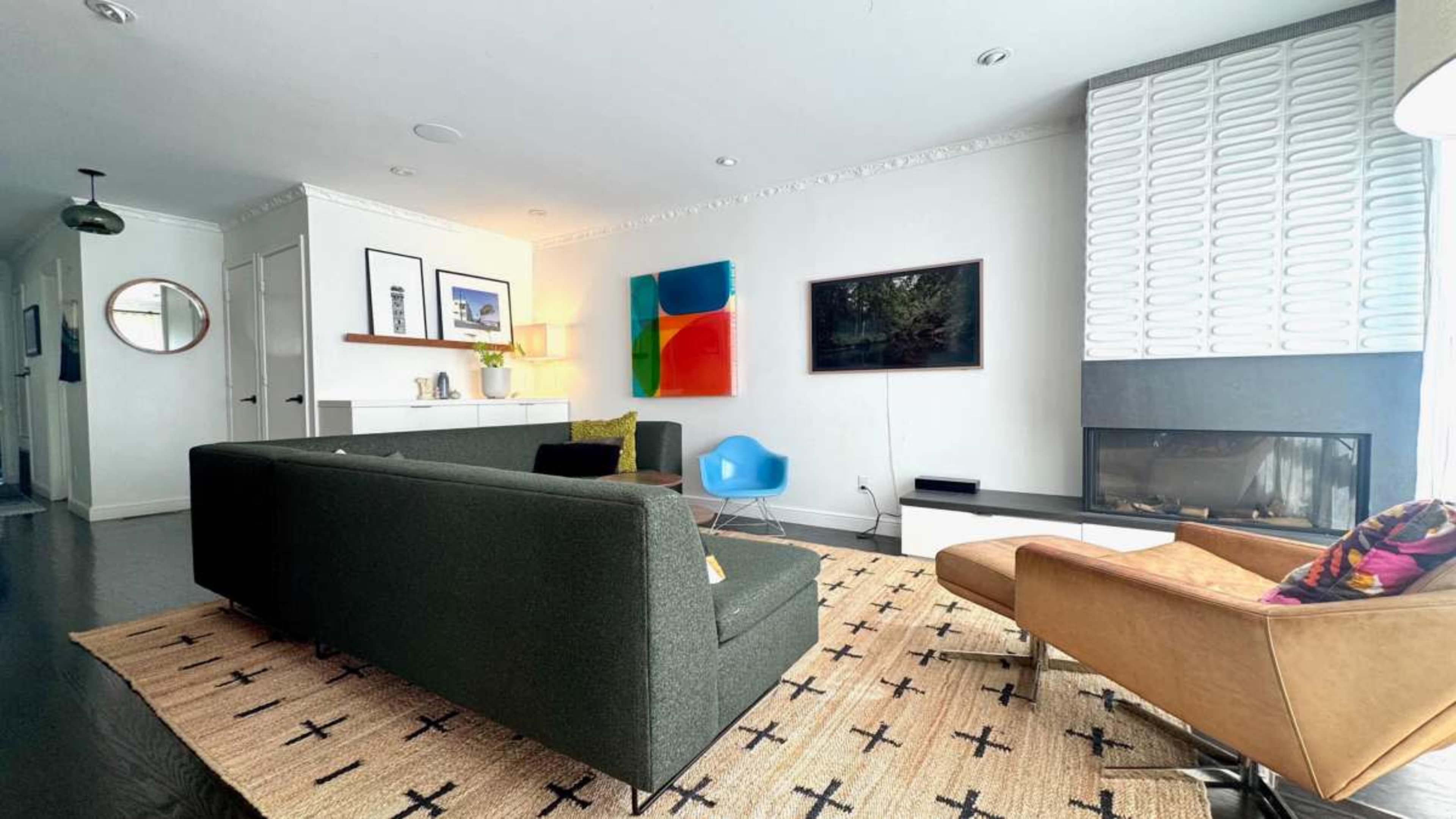 The living room features a modern design with a green sofa, a brown armchair, a low coffee table, and a large wall-mounted television, all arranged on a patterned rug.