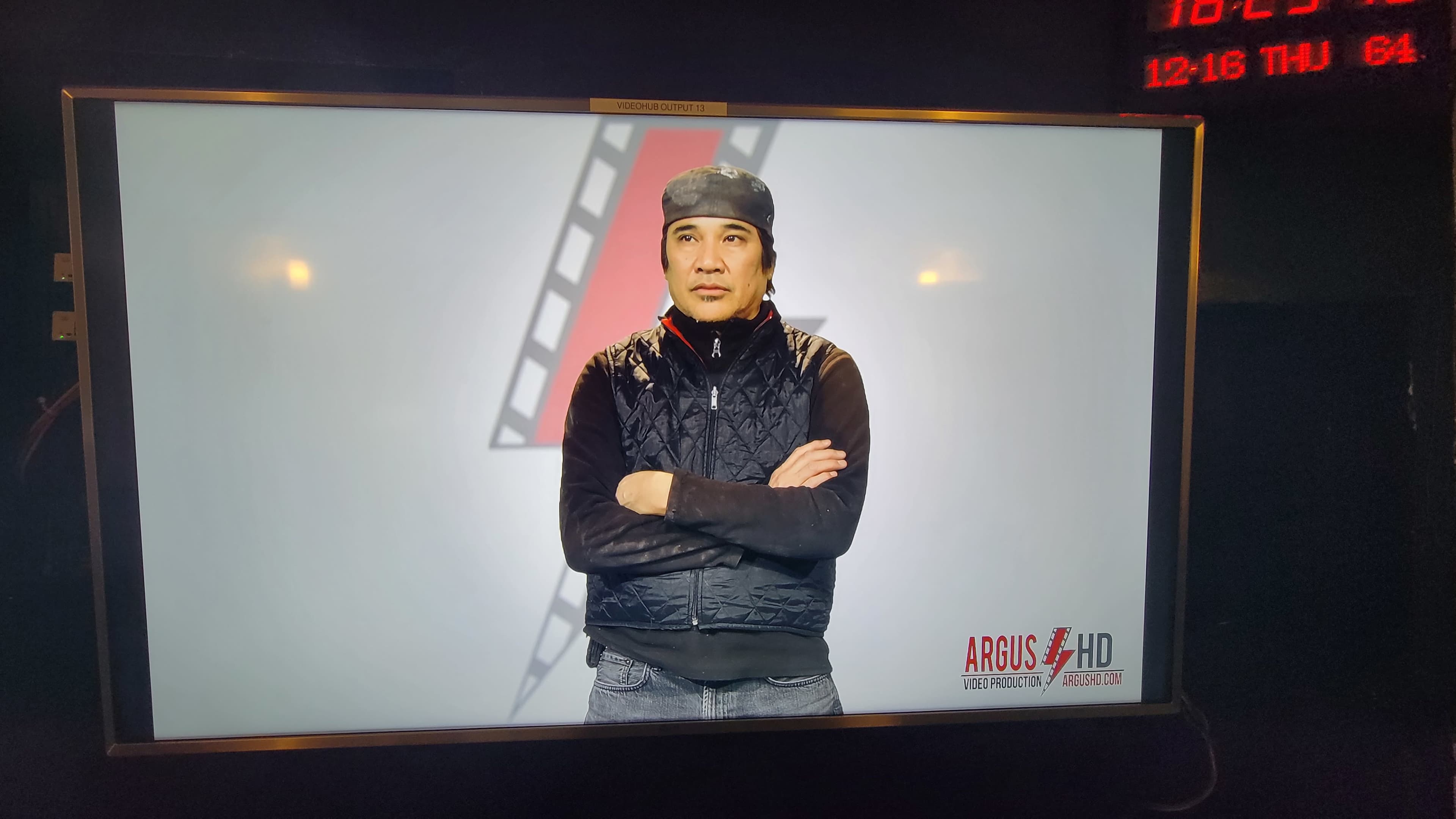 A man in a black vest and cap stands with his arms crossed on a screen displaying a movie-themed background.
