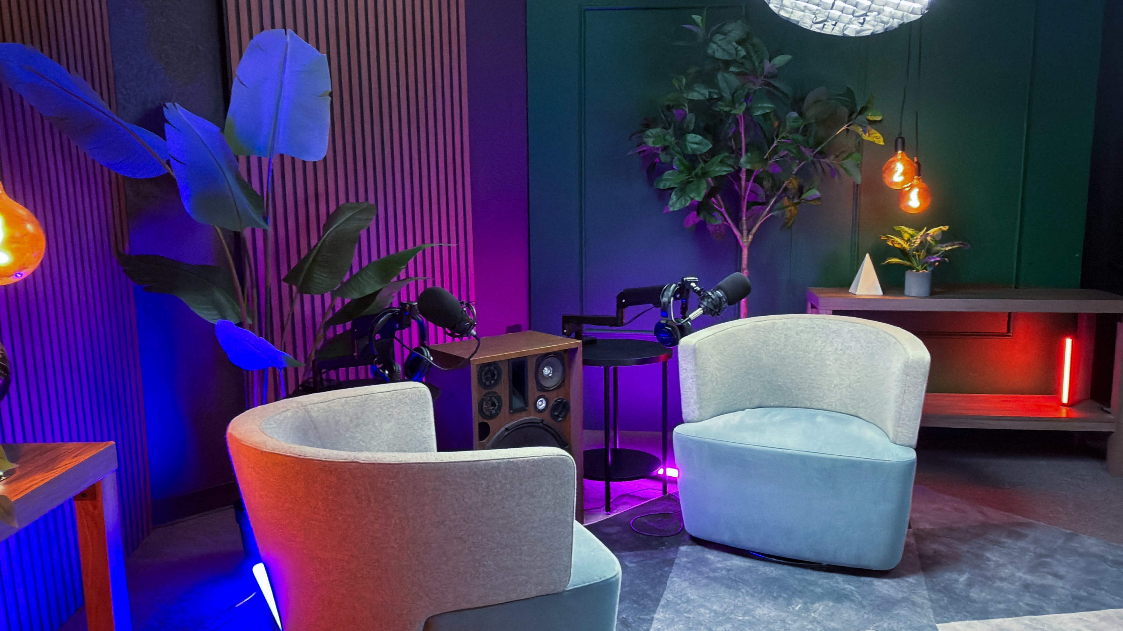 The image shows a modern, cozy recording studio setup with two plush chairs, microphones, and colorful lighting, surrounded by plants and decorative elements.