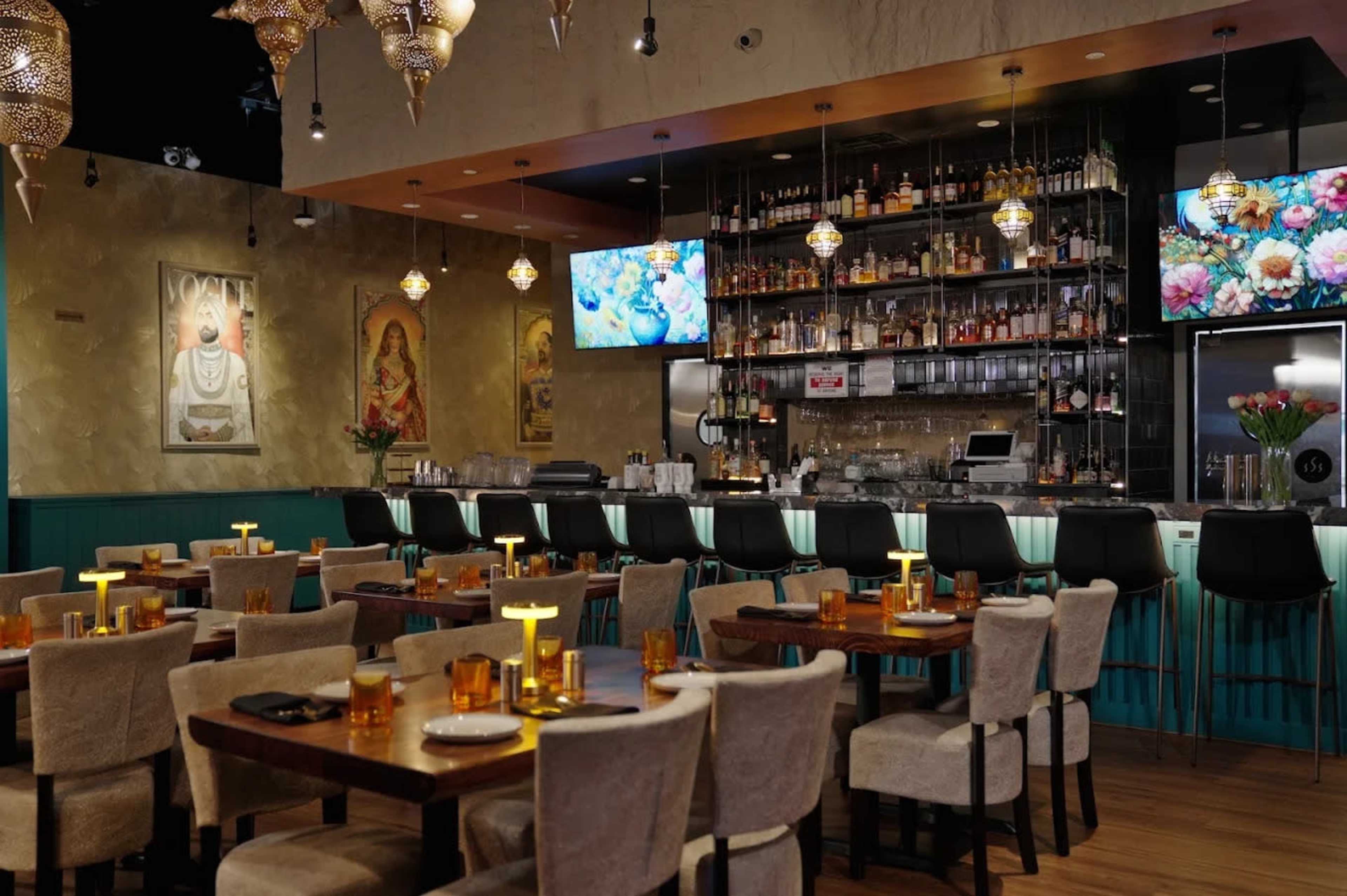 The image shows an elegantly designed restaurant interior featuring a bar in the background, wooden tables set for dining, and decorative lighting.