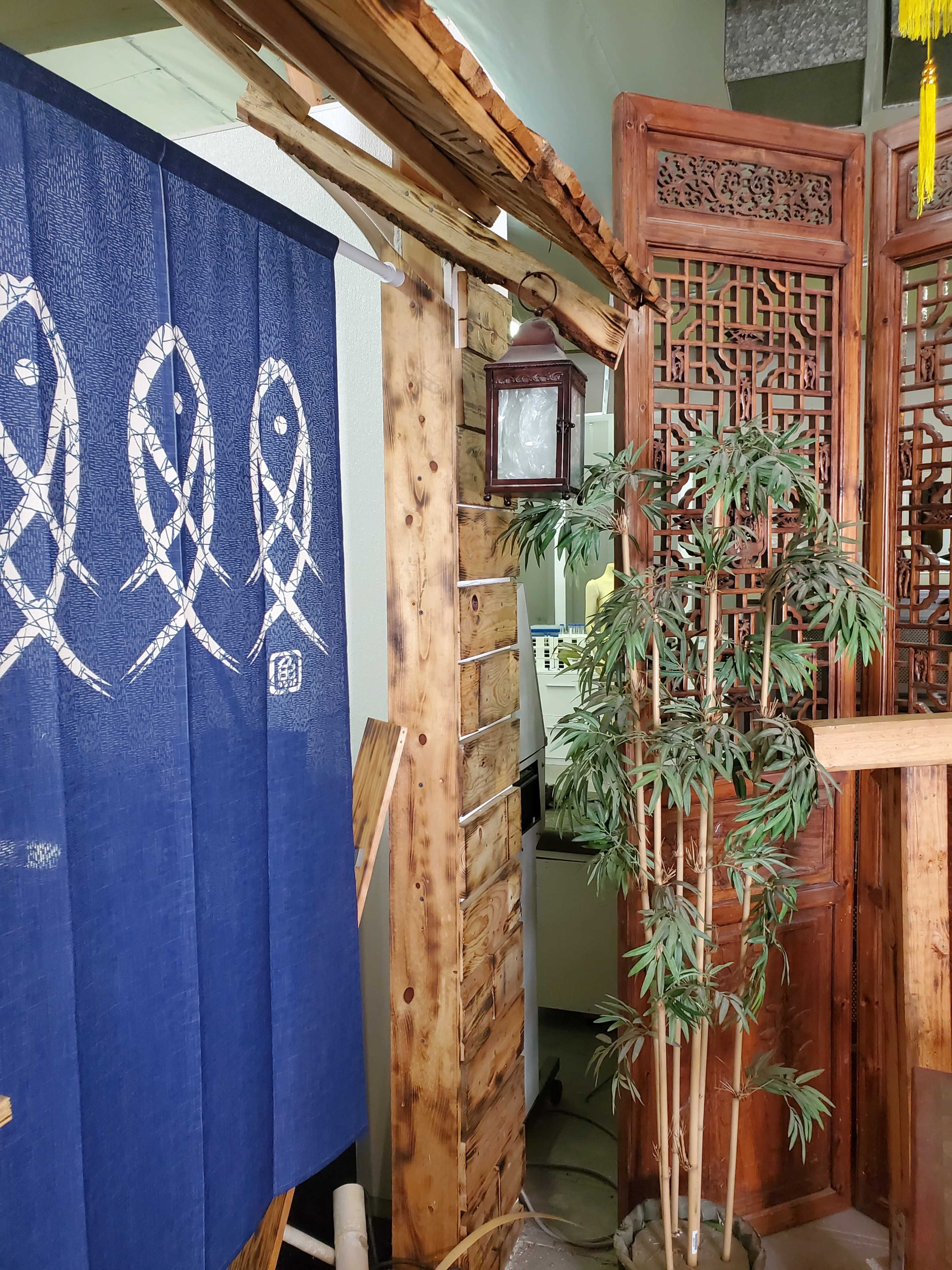 A wooden entrance featuring a blue fabric curtain with fish designs, flanked by decorative wooden screens and a potted plant.