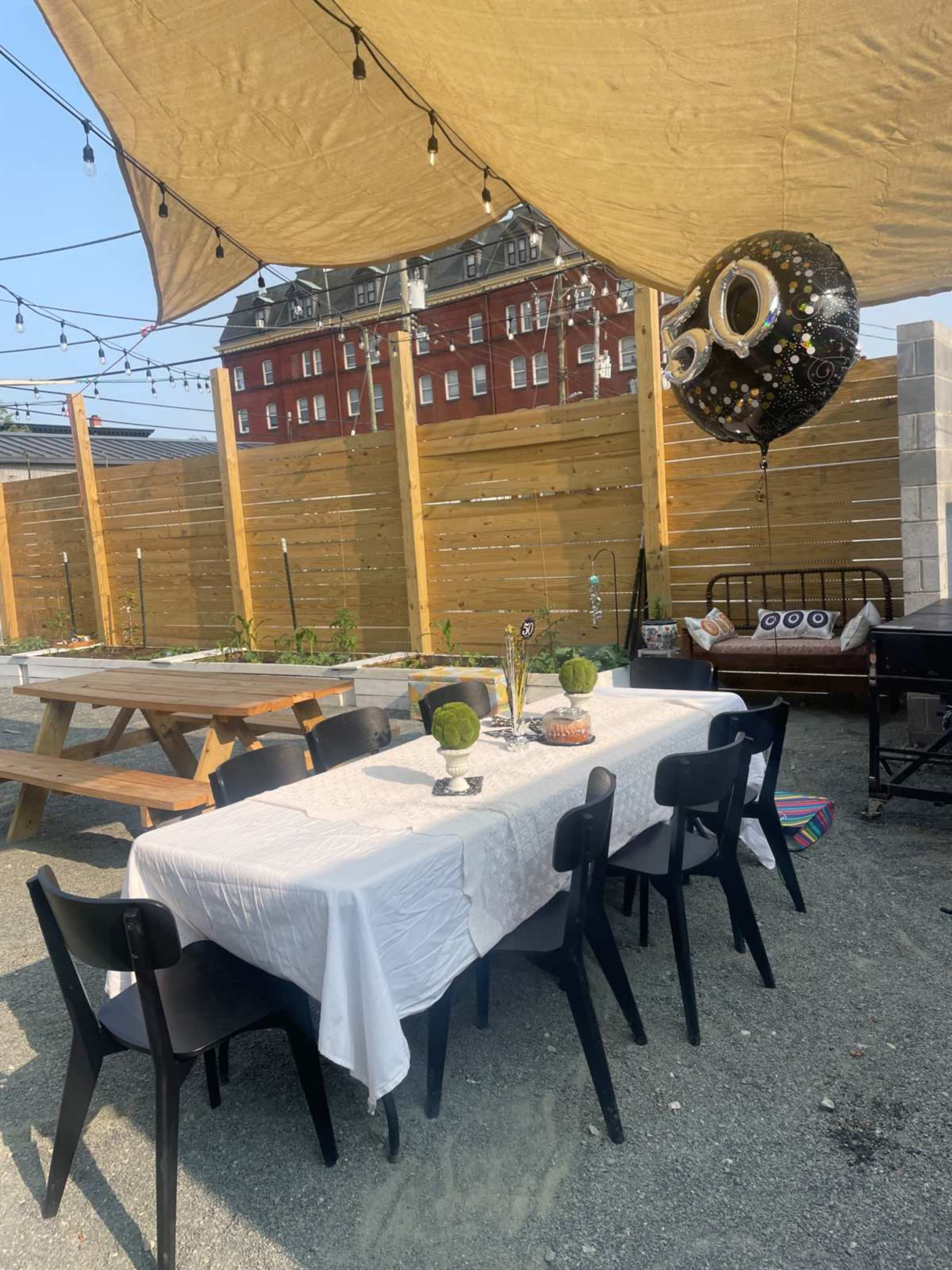 A large outdoor dining table with black chairs is set up under a canopy, adorned with a large balloon marked "50" and surrounded by decorative elements.