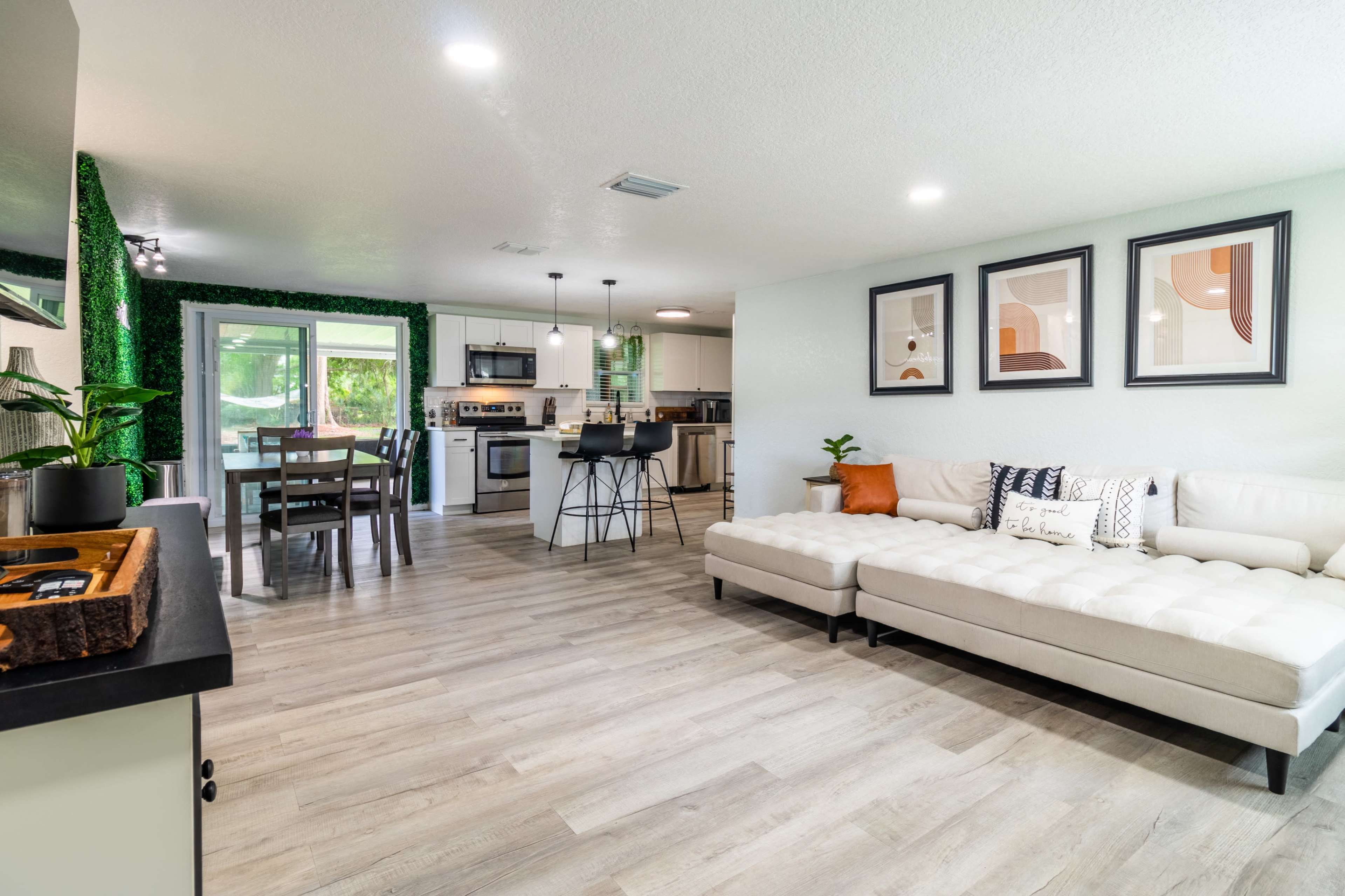 A modern open-concept living space featuring a white sectional sofa, a dining area with a wooden table, and a kitchen with stainless steel appliances.