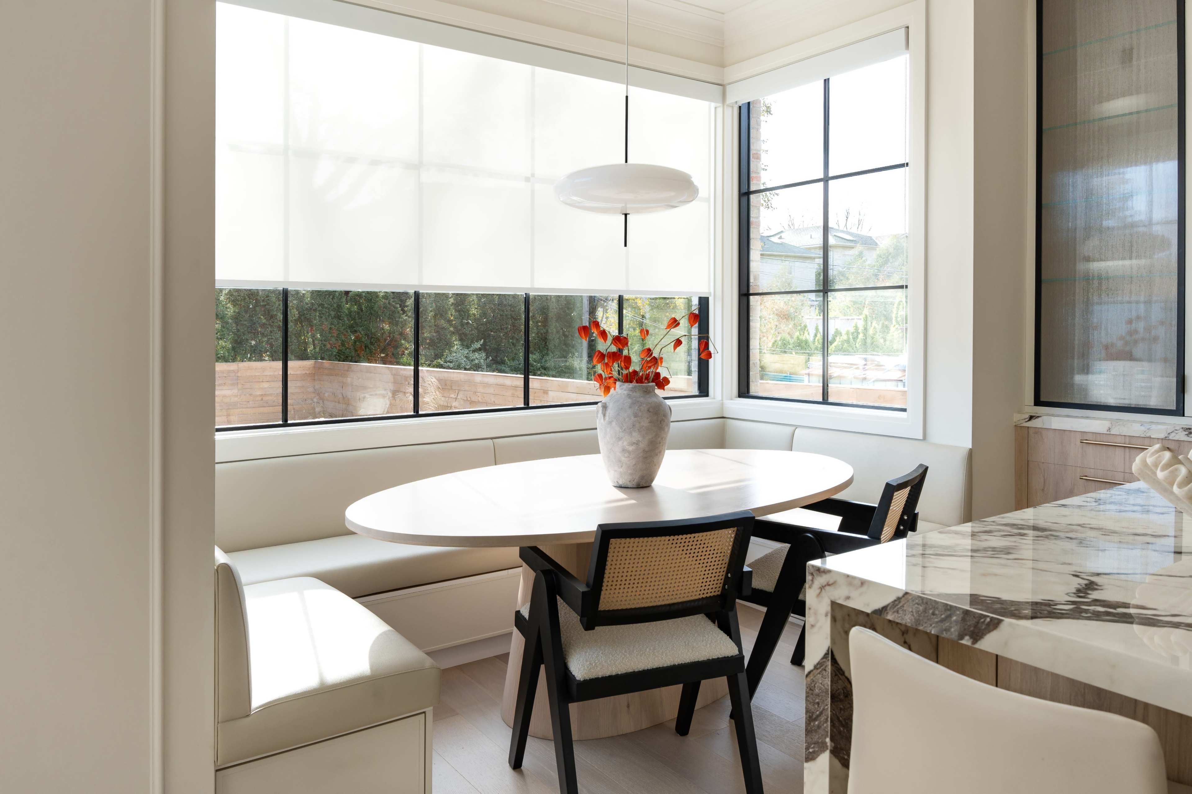 The image shows a modern dining area with an oval table, black and white chairs, and a vase of red flowers on the table, all illuminated by natural light from large windows.