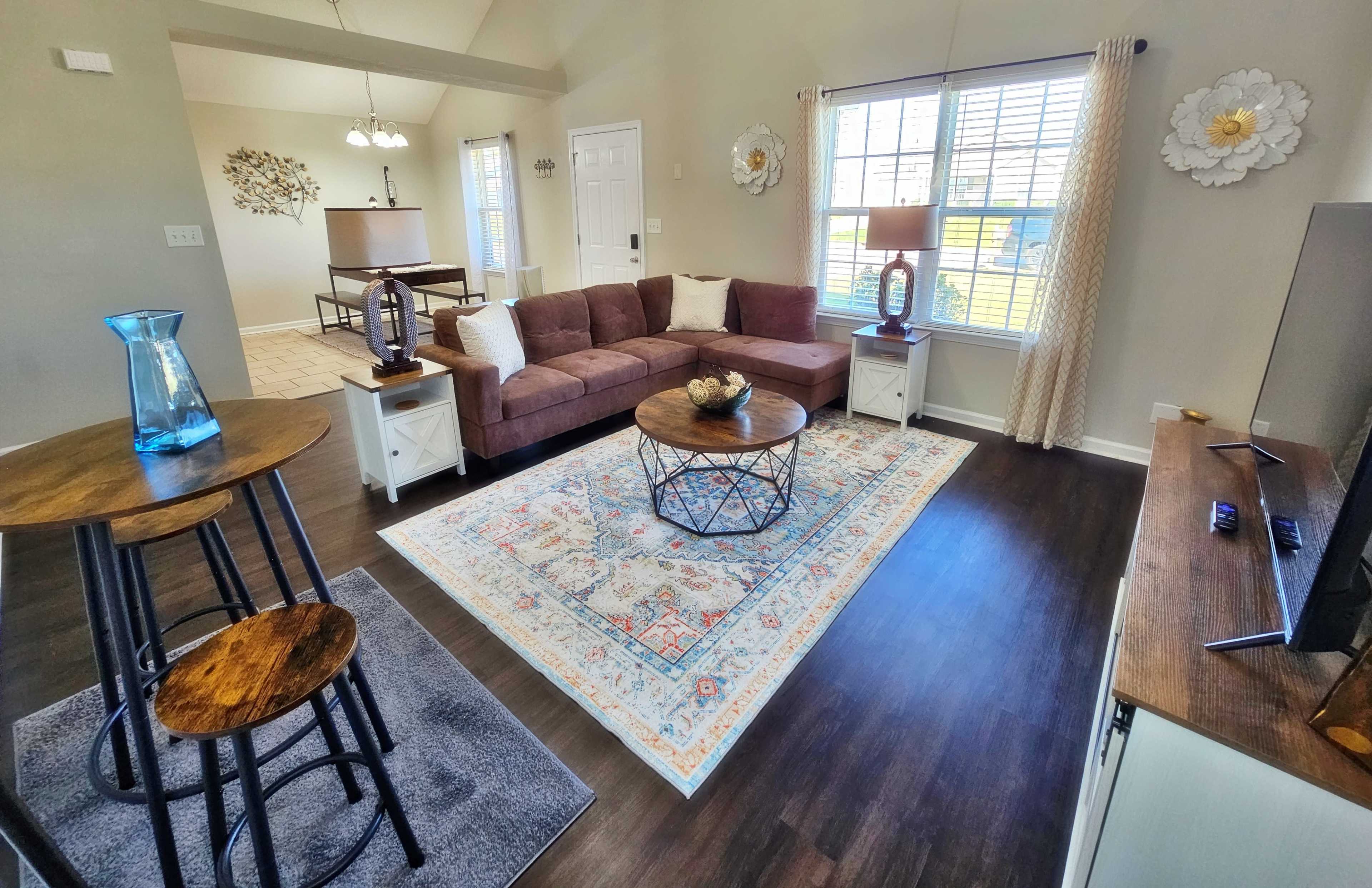The image shows a modern living room with a sectional sofa, a round coffee table on an area rug, and a small dining table with stools beside a window.