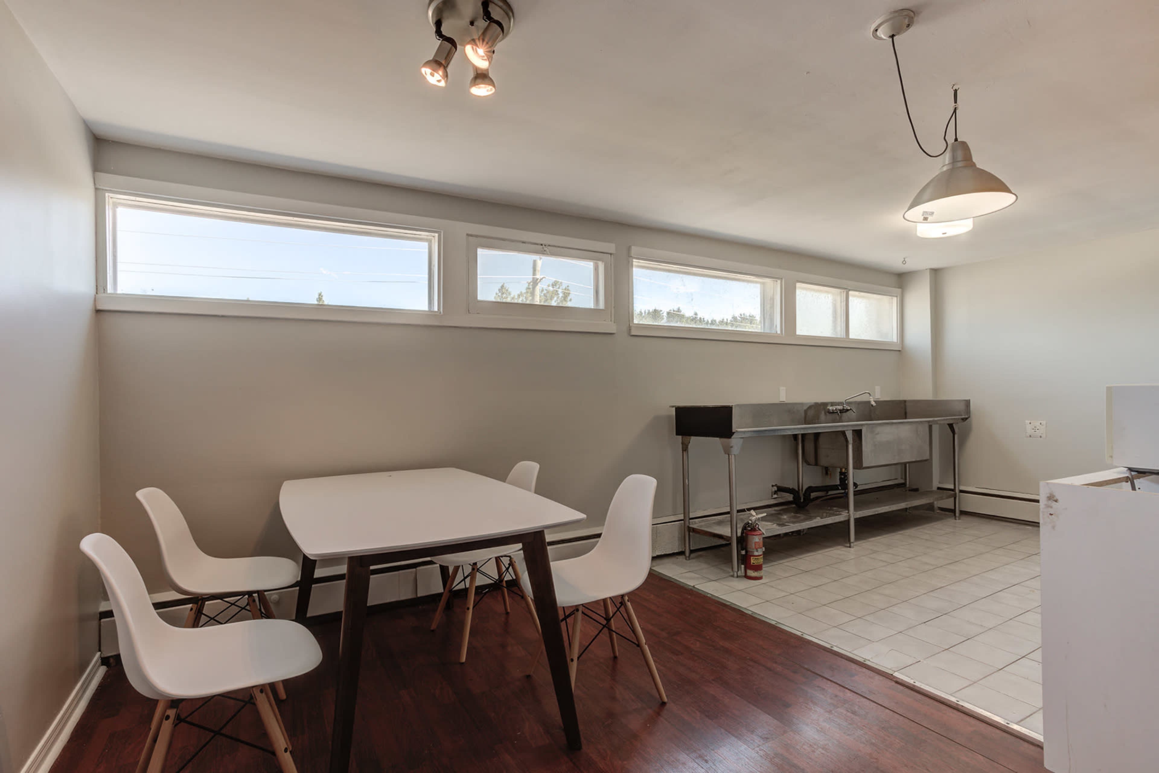 A minimalist kitchen area featuring a table with four chairs, a stainless steel sink, and several windows allowing natural light.