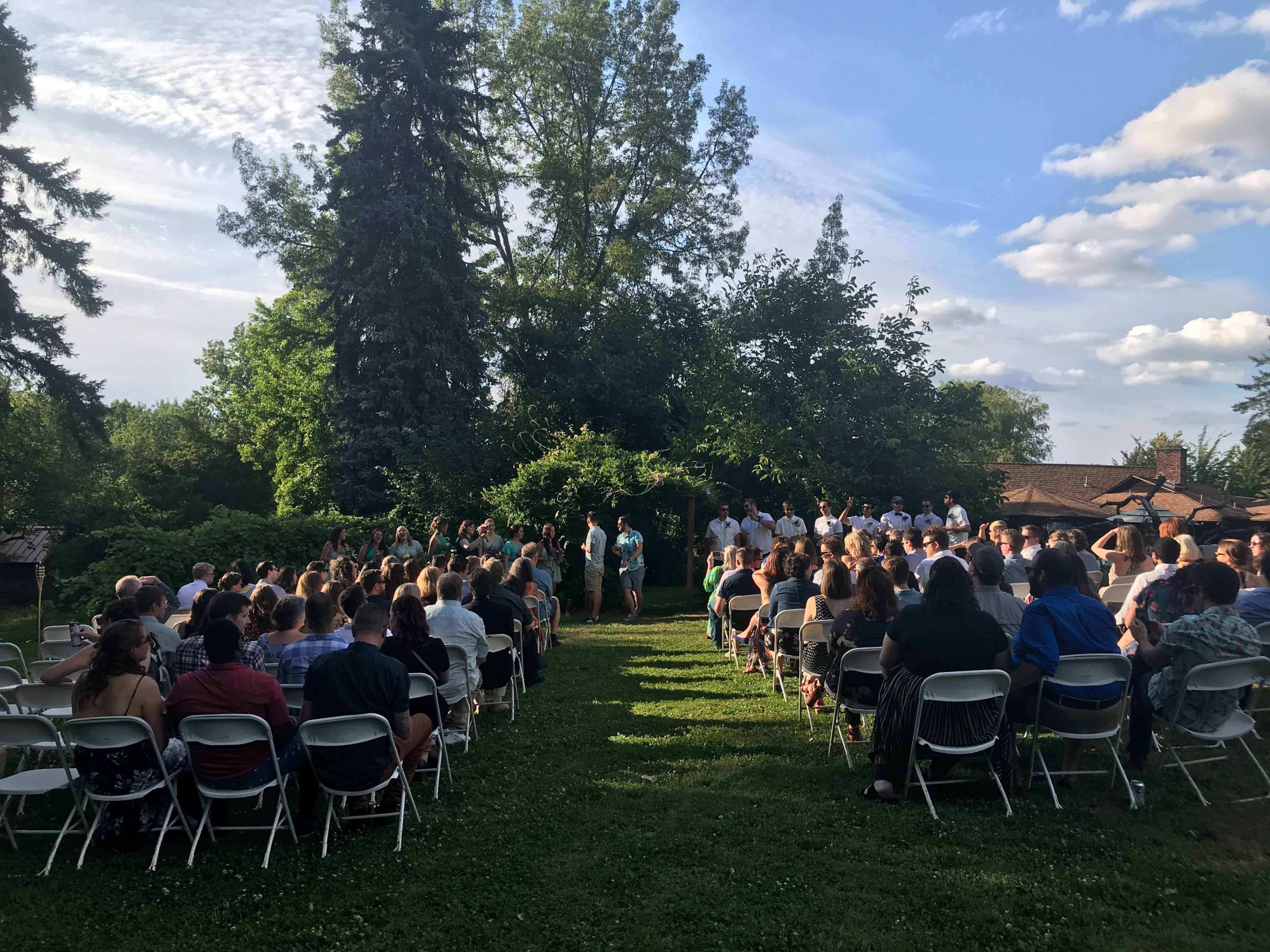 A large group of people is gathered on chairs in a grassy area for a wedding ceremony, with trees and a blue sky in the background.