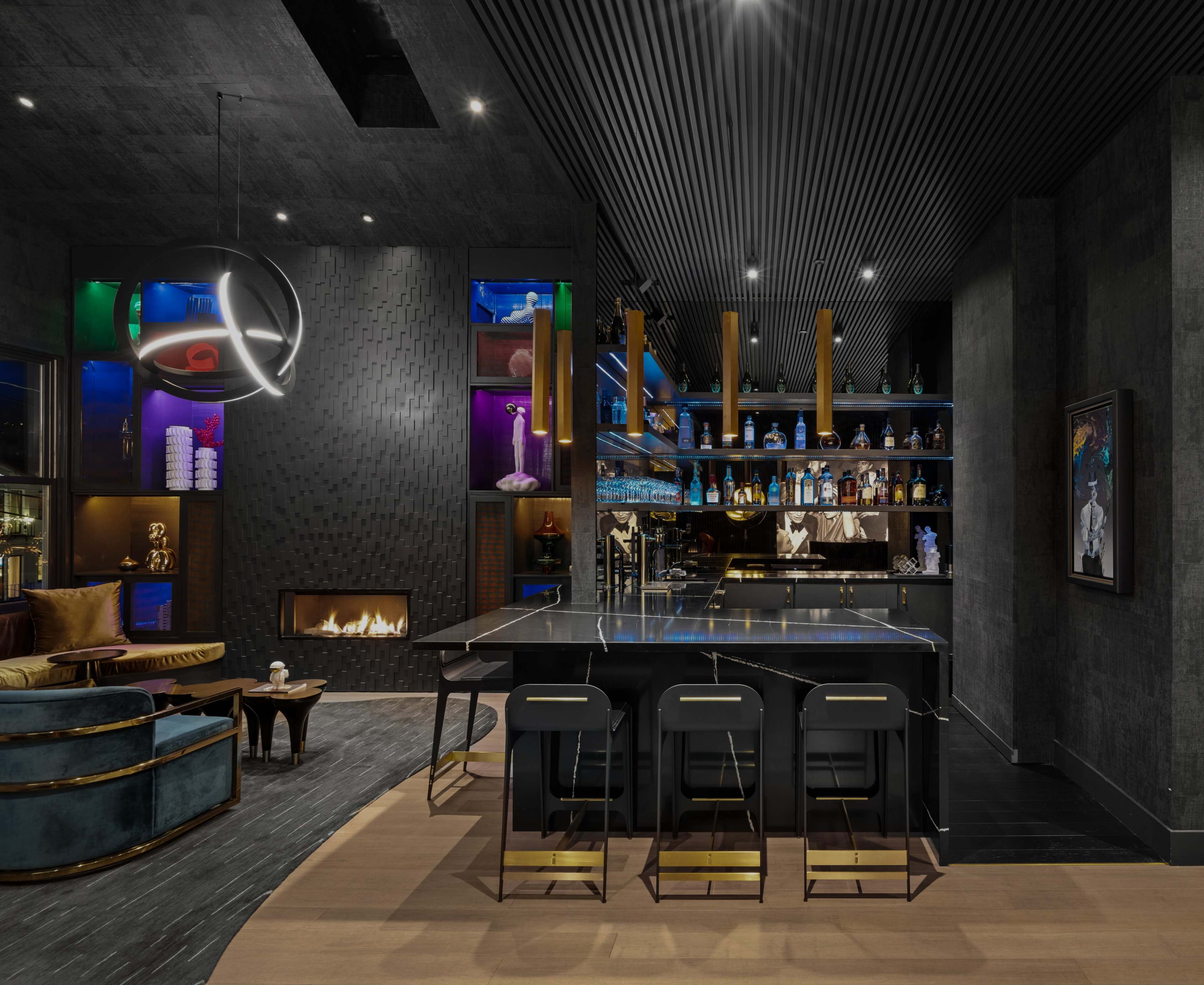 A modern bar area features a sleek black countertop, illuminated shelves displaying various bottles, and a cozy seating arrangement with a fireplace.