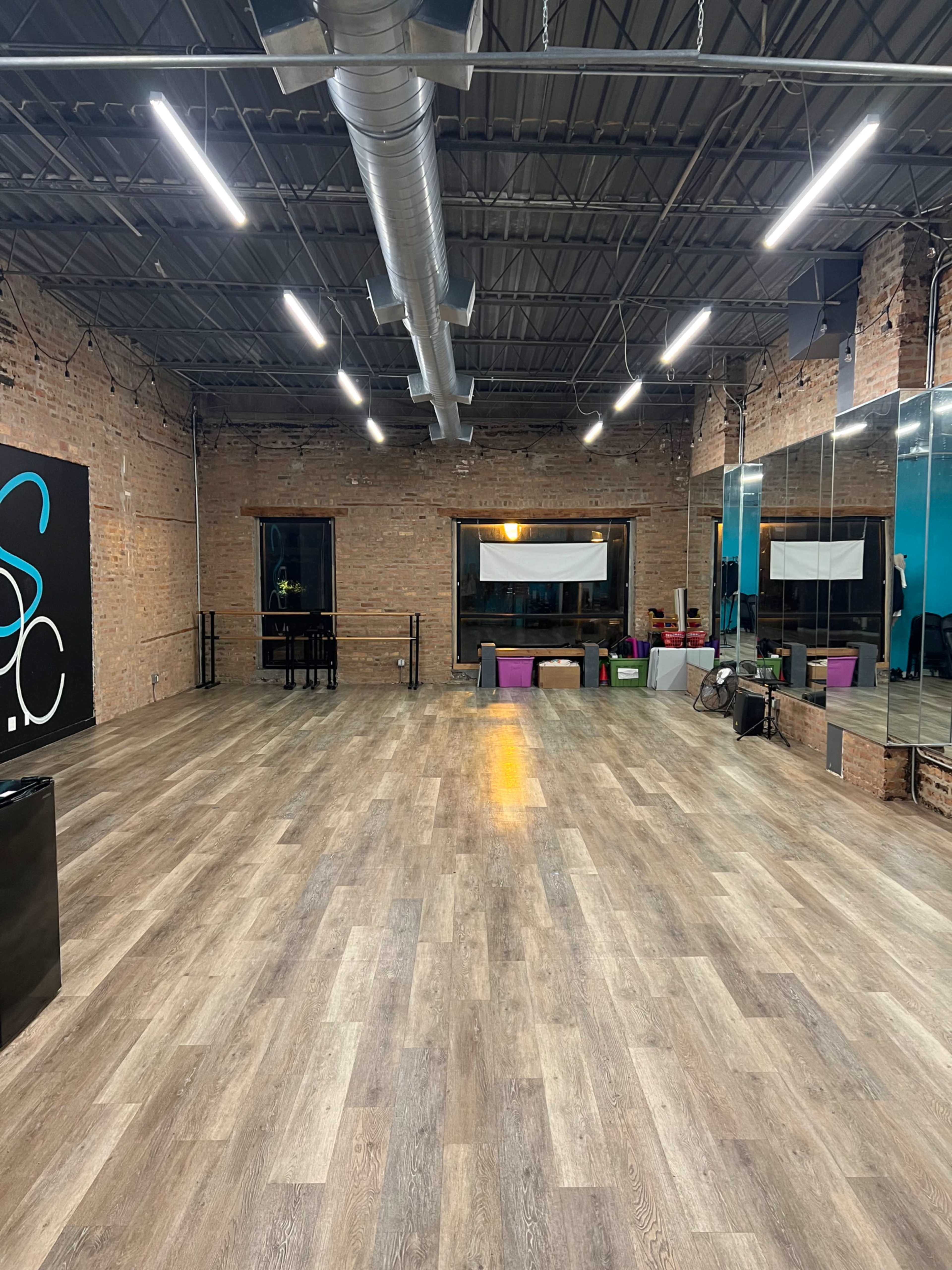 A spacious, well-lit dance studio with wooden flooring, exposed brick walls, and mirrors along one side.