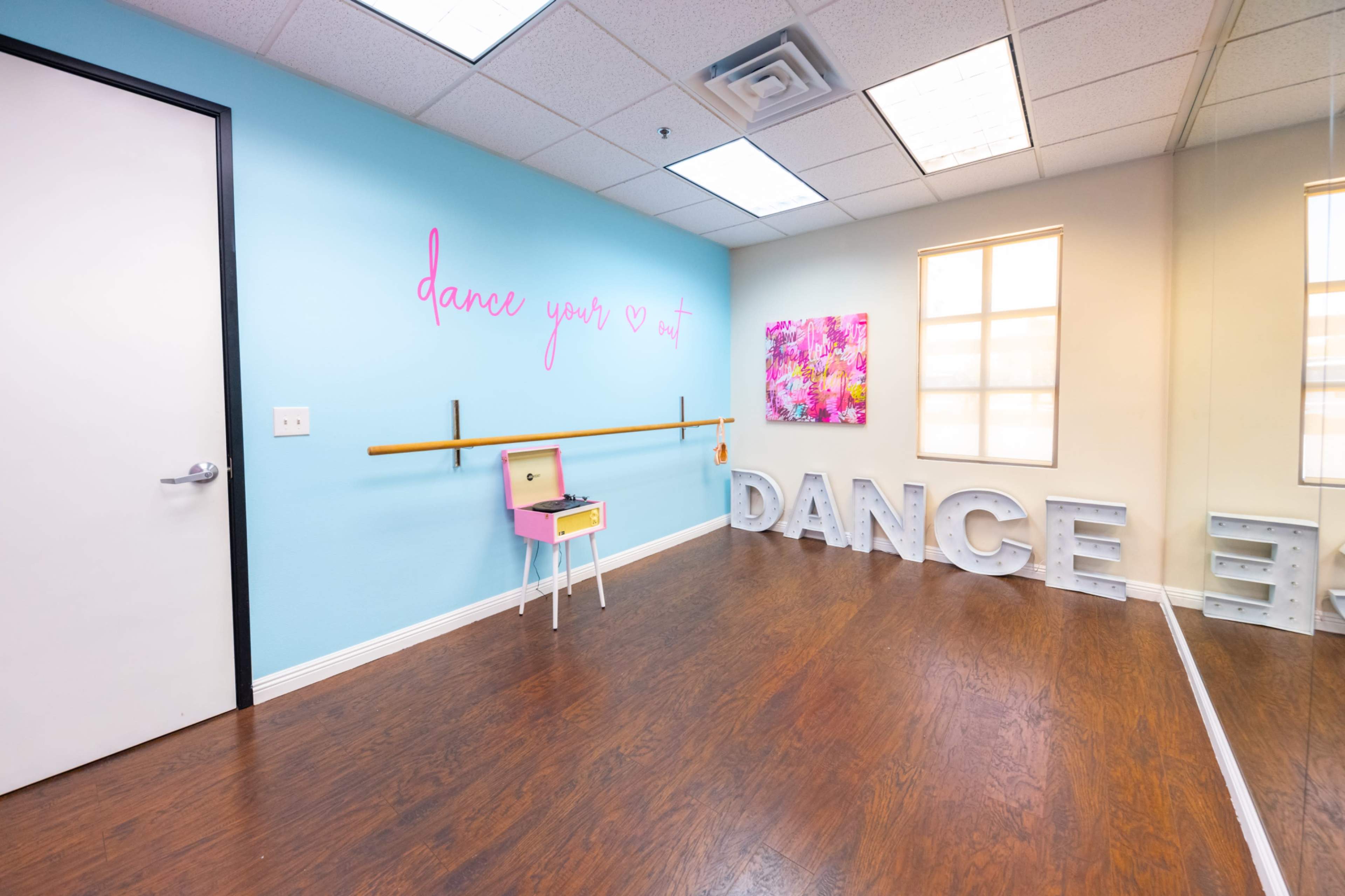 A dance studio features a mirrored wall, a barre, and colorful decor, including a wall with the phrase "dance your heart out."