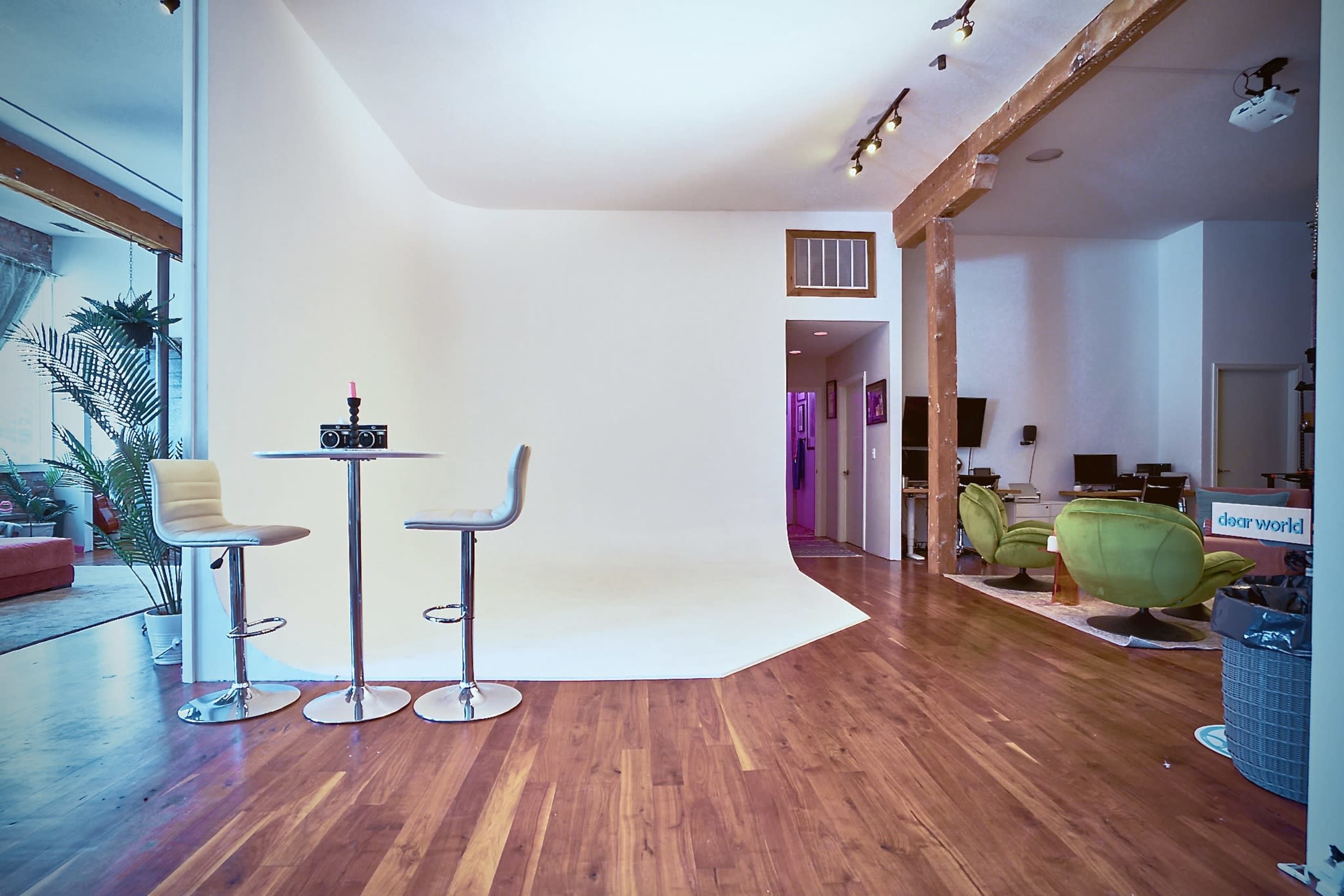 A spacious room with a white backdrop, two modern bar stools at a round table, and distinct areas featuring green chairs and various decor.