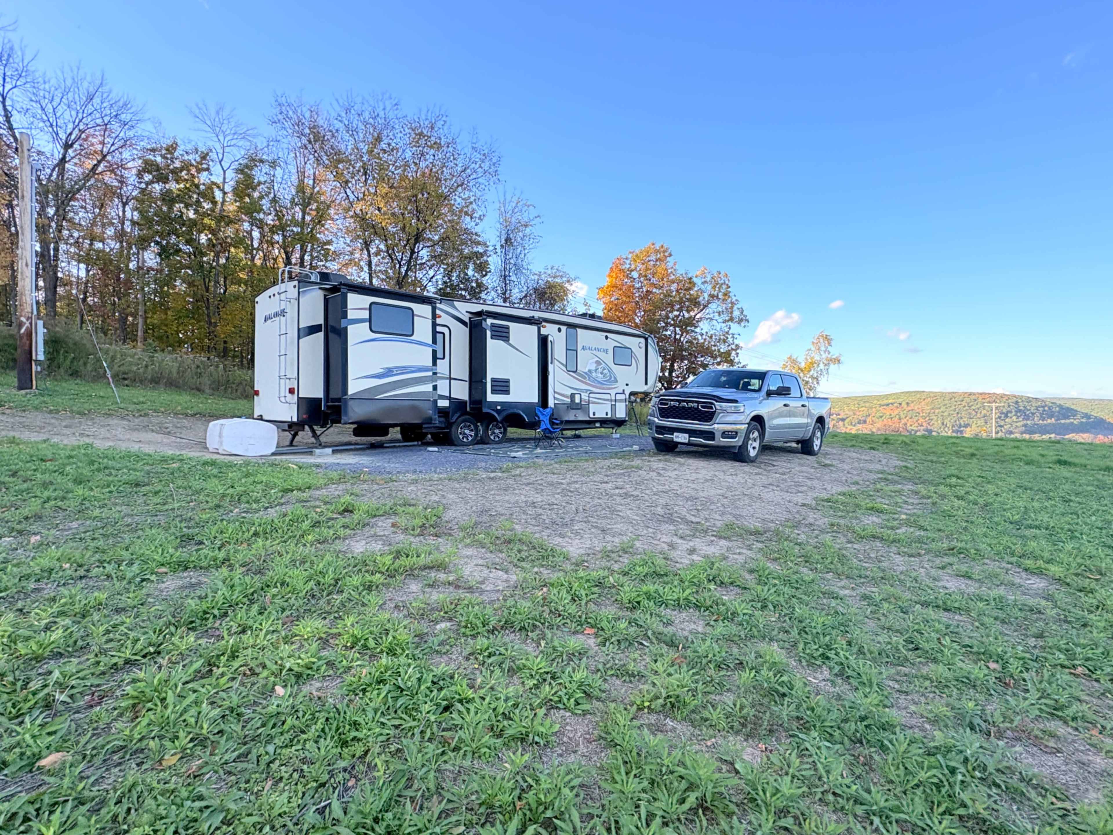 A recreational vehicle is parked next to a pickup truck on a grassy area with trees and hills in the background.