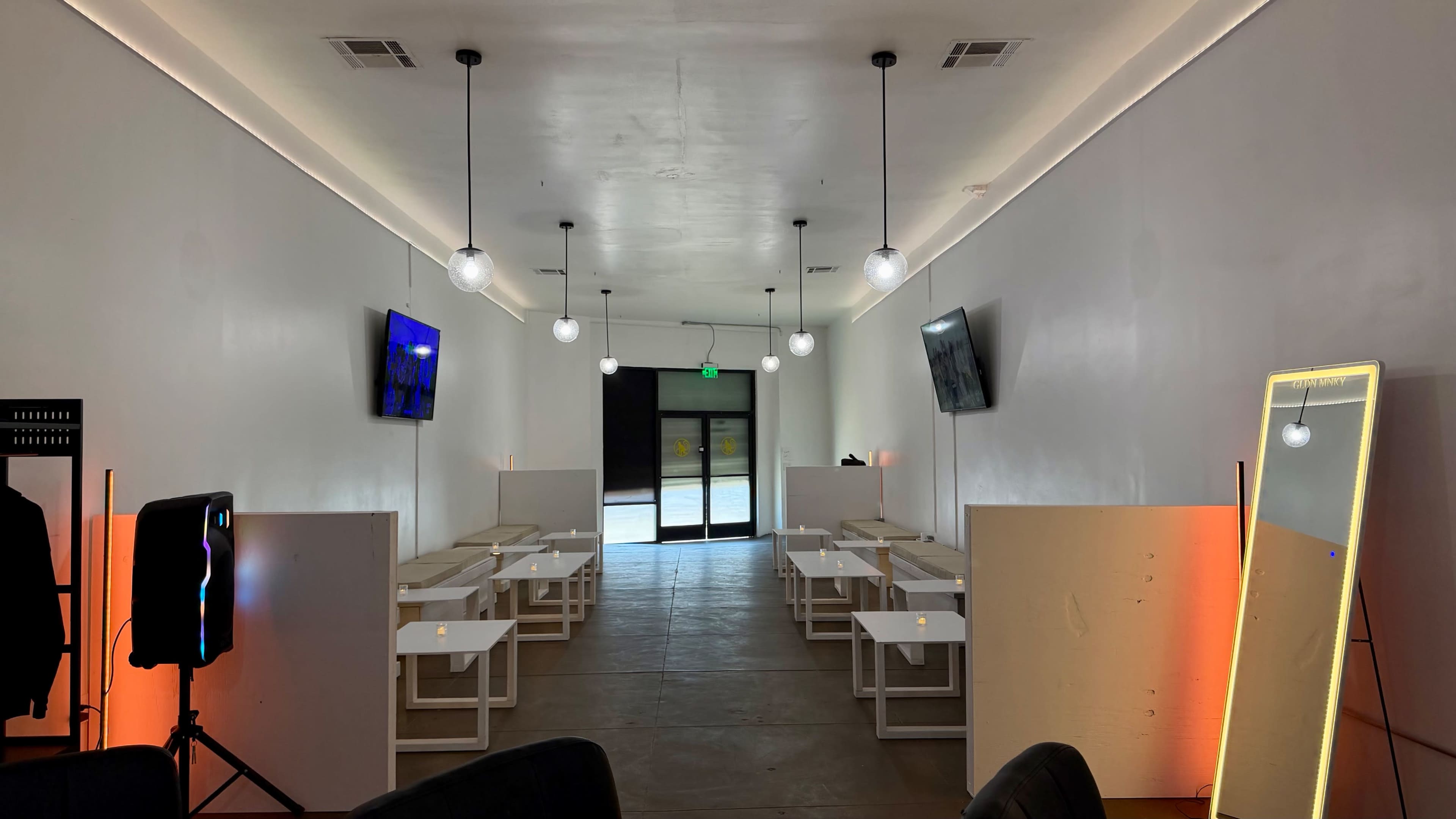 The image shows a narrow, well-lit interior space with several white tables arranged in rows, two televisions on the walls, and a large glass door at the far end.