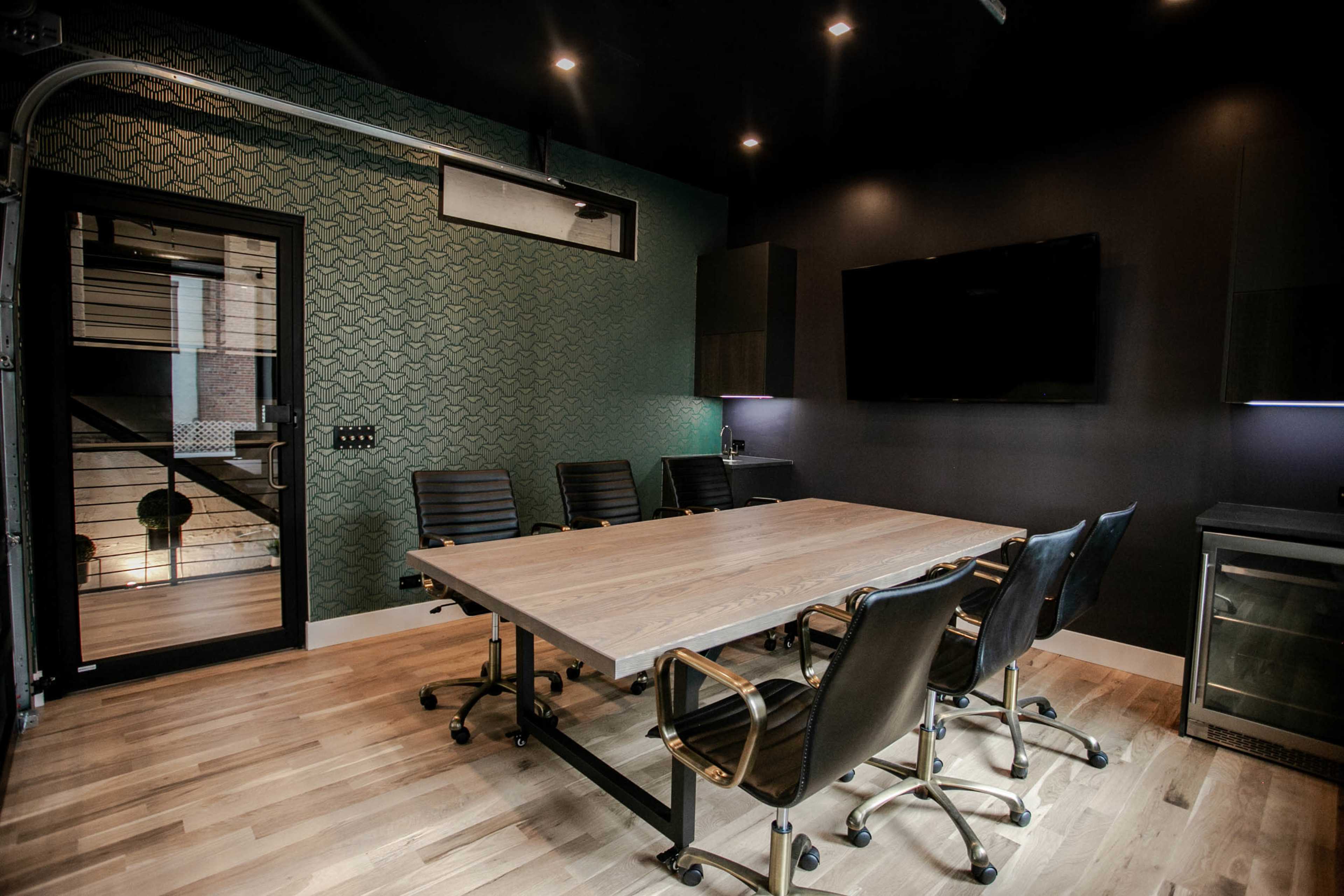 A modern conference room features a large wooden table, black ergonomic chairs, and a glass door leading to a staircase.