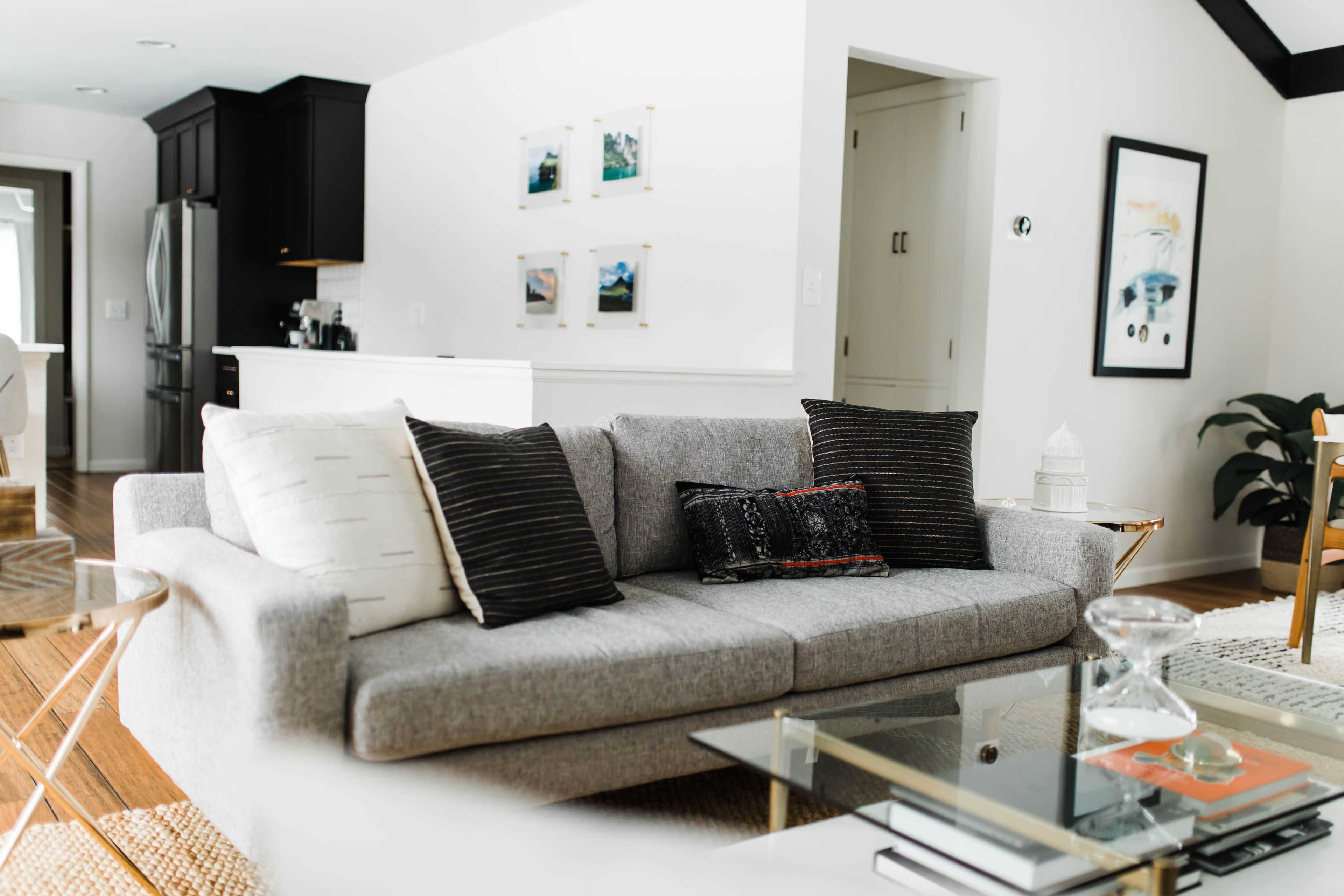 A gray sofa with patterned pillows is positioned in a modern living room, featuring a glass coffee table and white walls adorned with framed artwork.