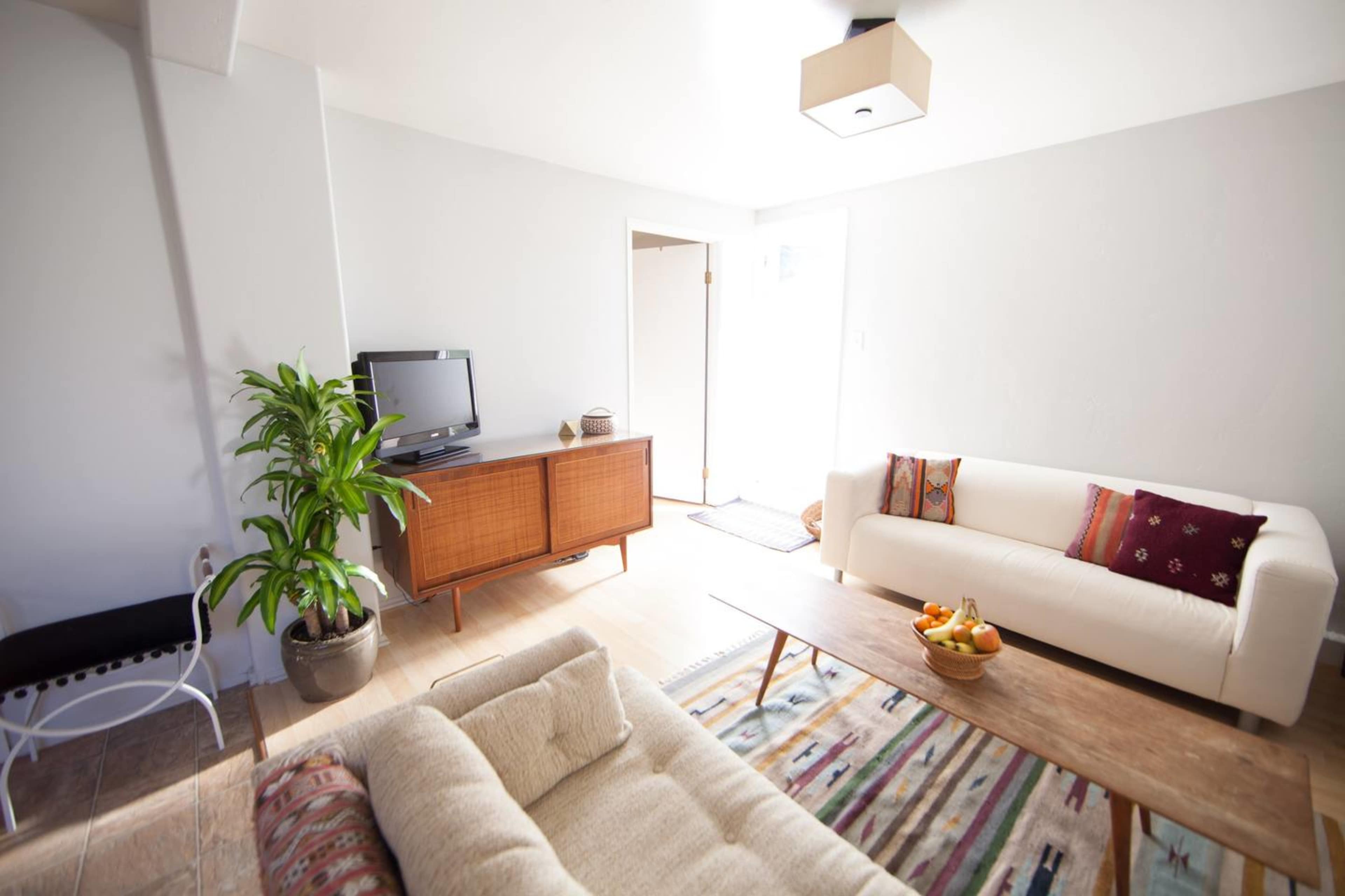 The image shows a light-colored living room with a white sofa, a wooden coffee table, a television on a cabinet, and a decorative plant in the corner.