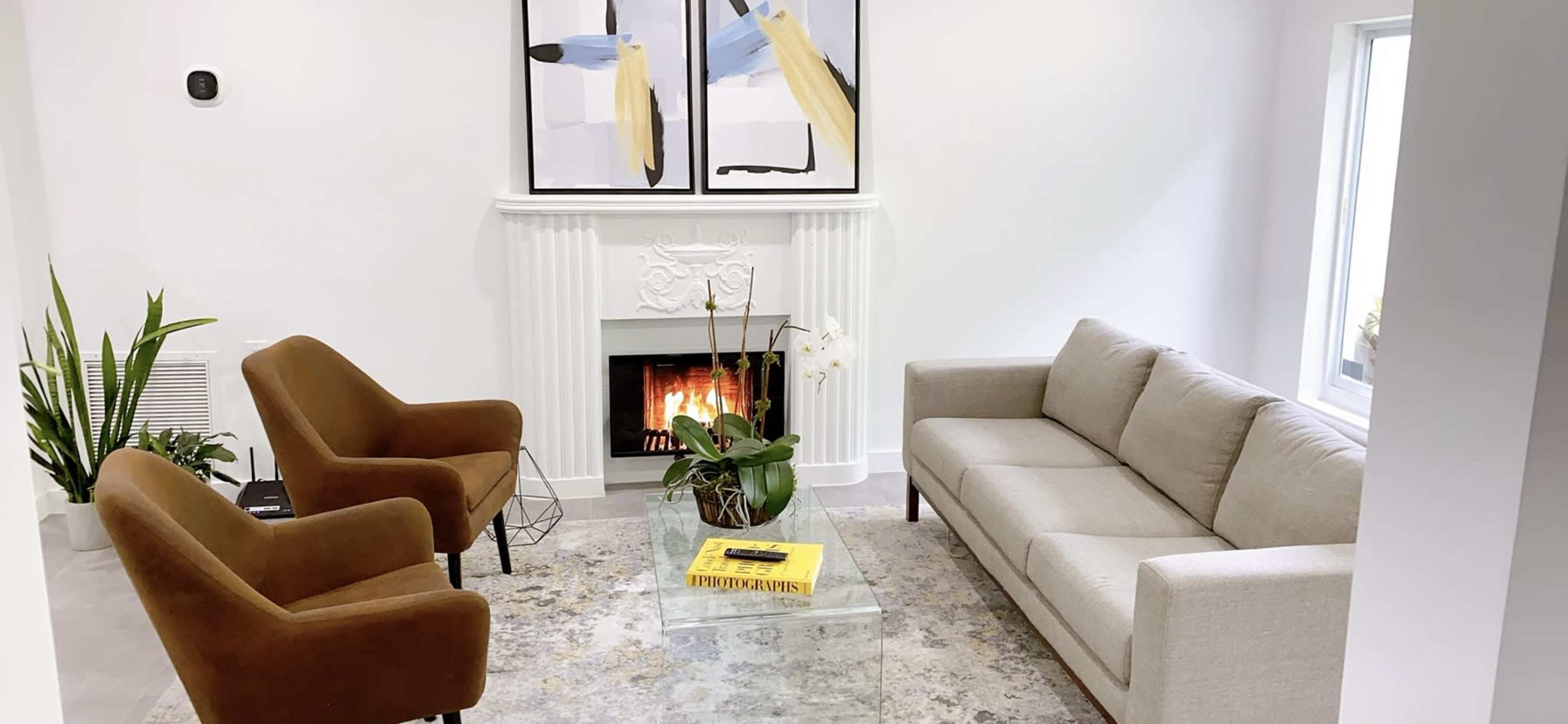 A modern living room with a beige sofa, two brown armchairs, a glass coffee table, a decorative plant, and a fireplace with a mantel, adorned with abstract art.