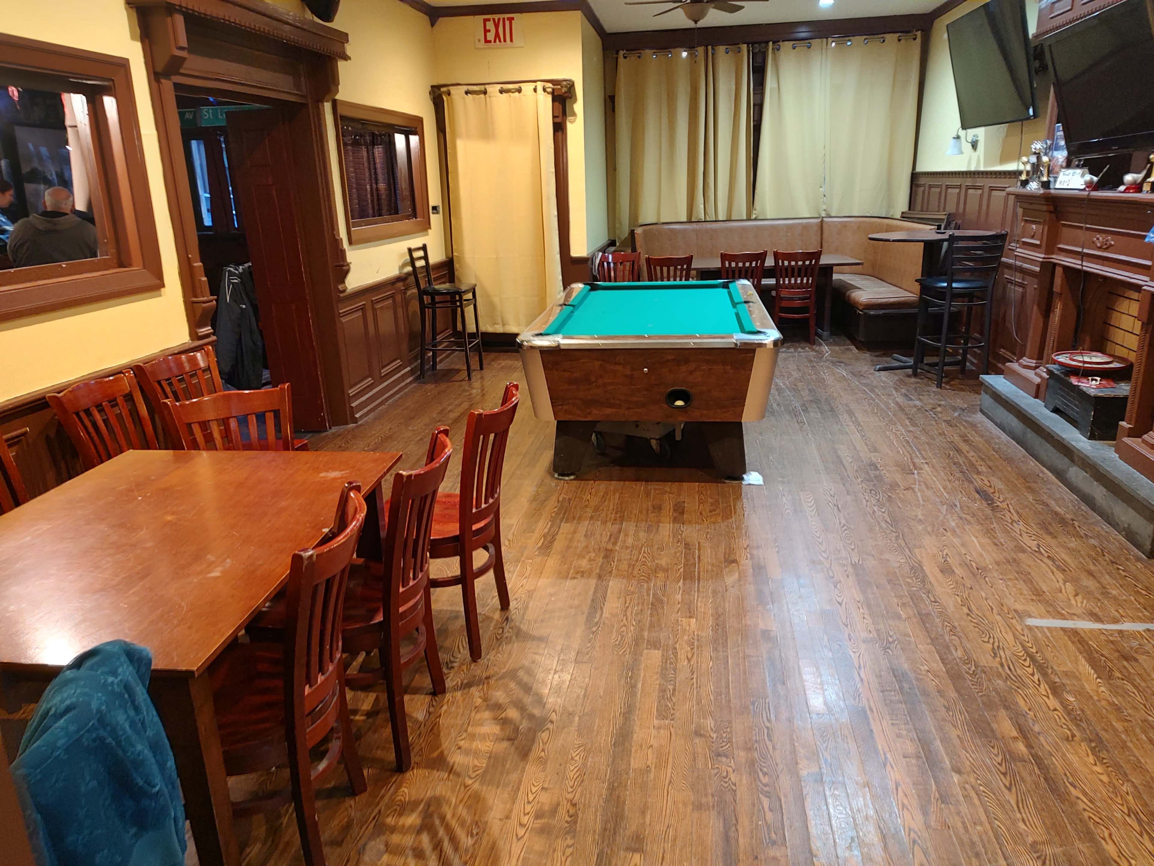 The image shows a rustic interior of a bar or pub, featuring wooden tables and chairs, a pool table in the center, and a small bar area along one wall.