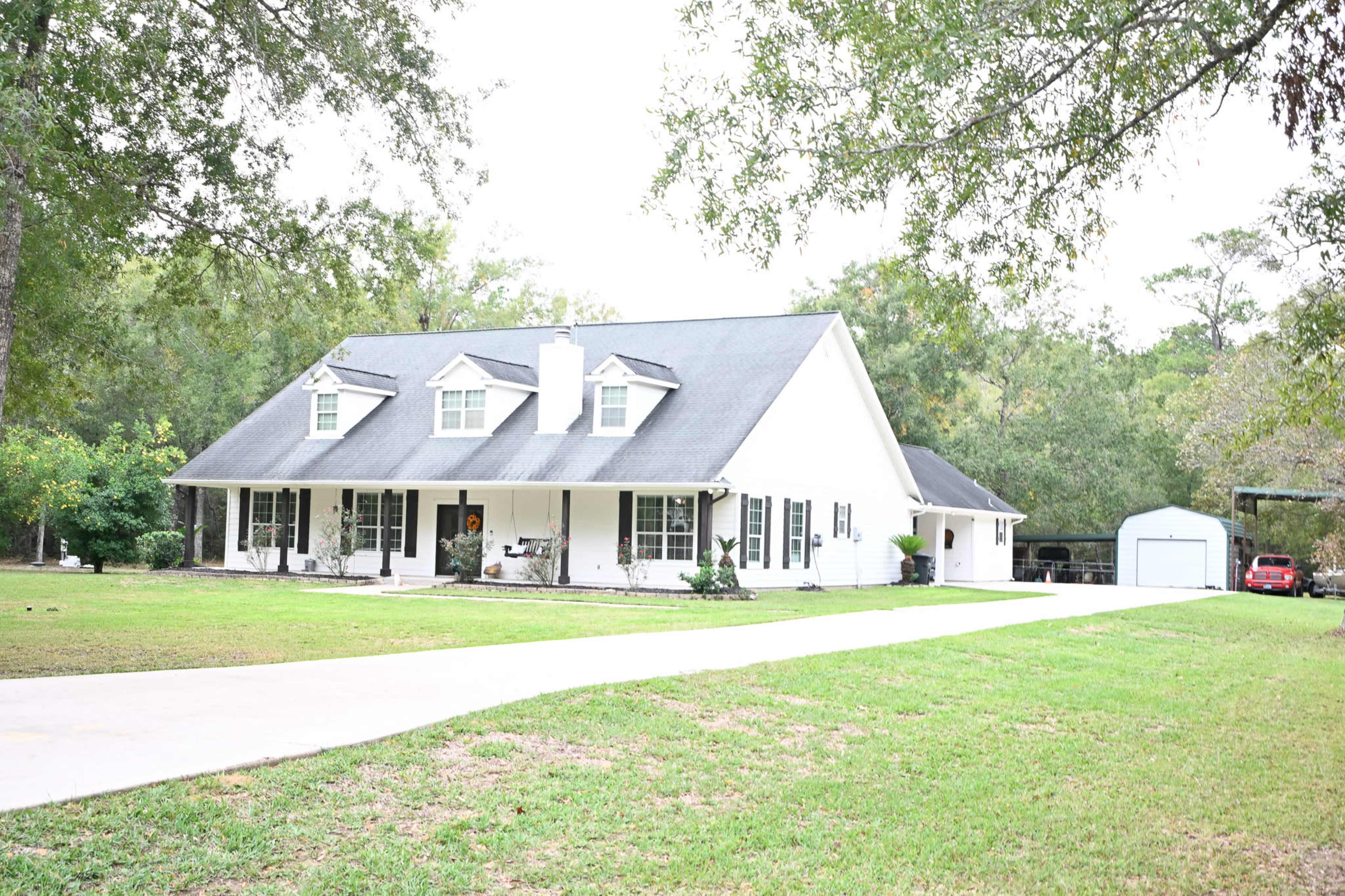 A single-story white house with a black roof is set on a spacious lawn, featuring a long driveway and a nearby outbuilding.