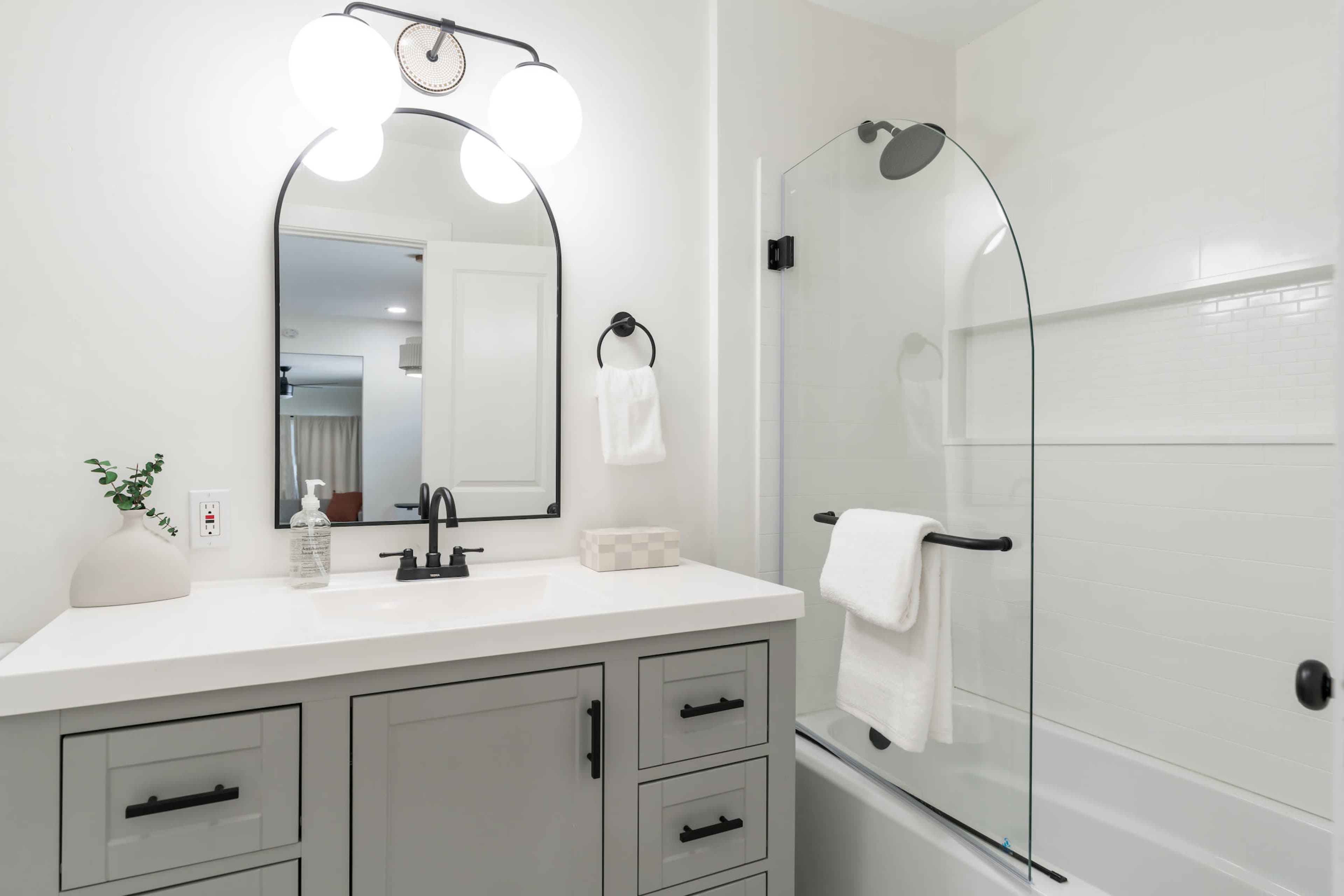 The image shows a modern bathroom featuring a gray vanity with a sink, a large mirror with rounded edges, and a glass shower enclosure beside a bathtub.