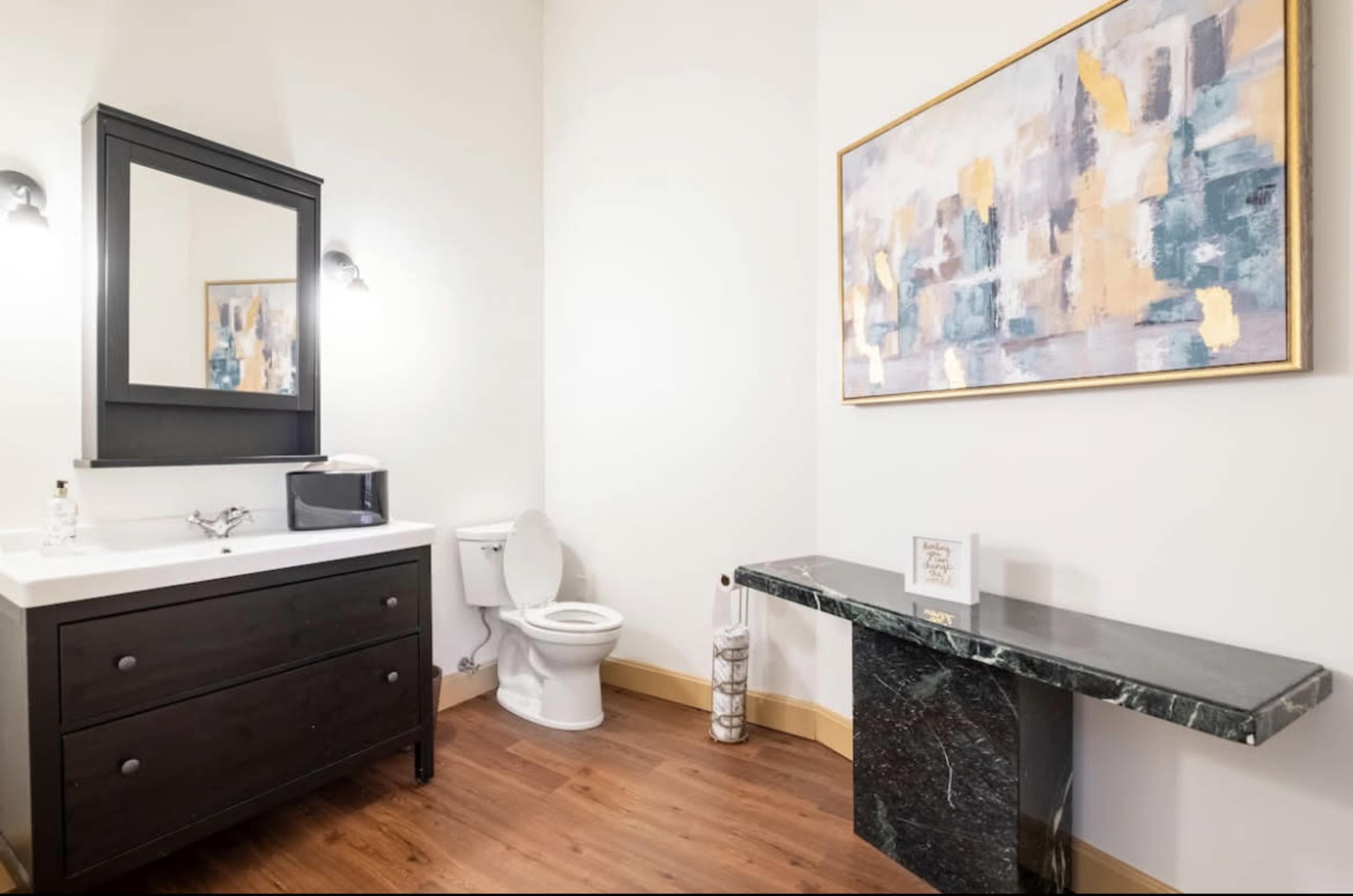 A modern bathroom with a dark wooden vanity, a white sink, a toilet, and a marble-topped console table next to a colorful abstract painting on the wall.