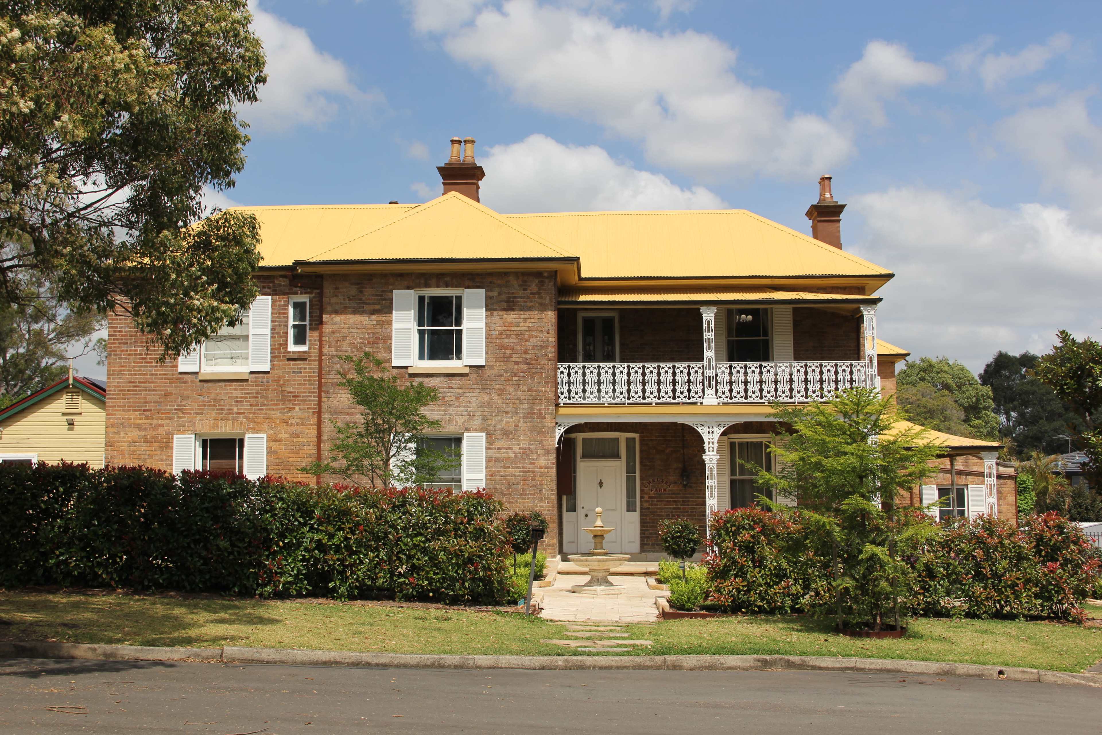 The image shows a two-story brick house with a yellow roof and a front porch, surrounded by well-maintained gardens and shrubs.