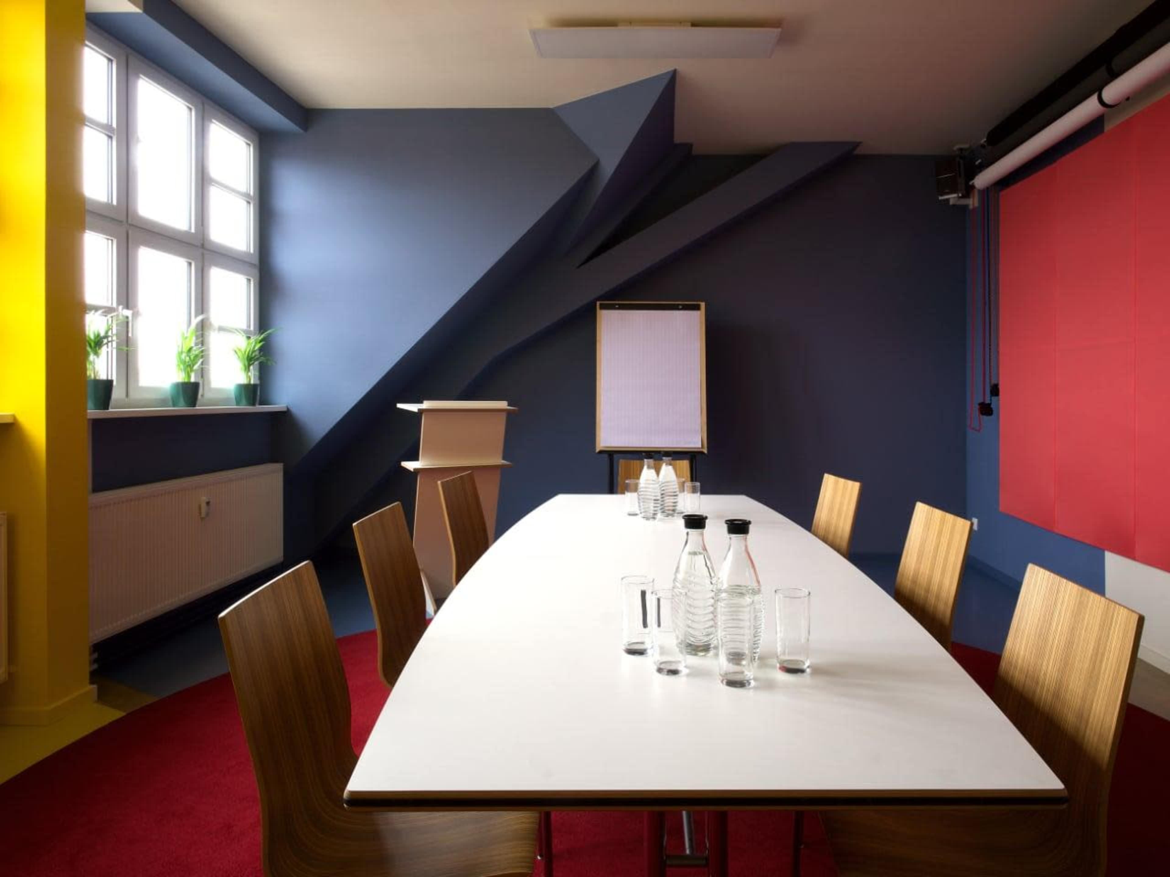 A modern conference room features a long white table surrounded by wooden chairs, with a flip chart and glass water bottles positioned on the table against a backdrop of colorful walls and large windows.