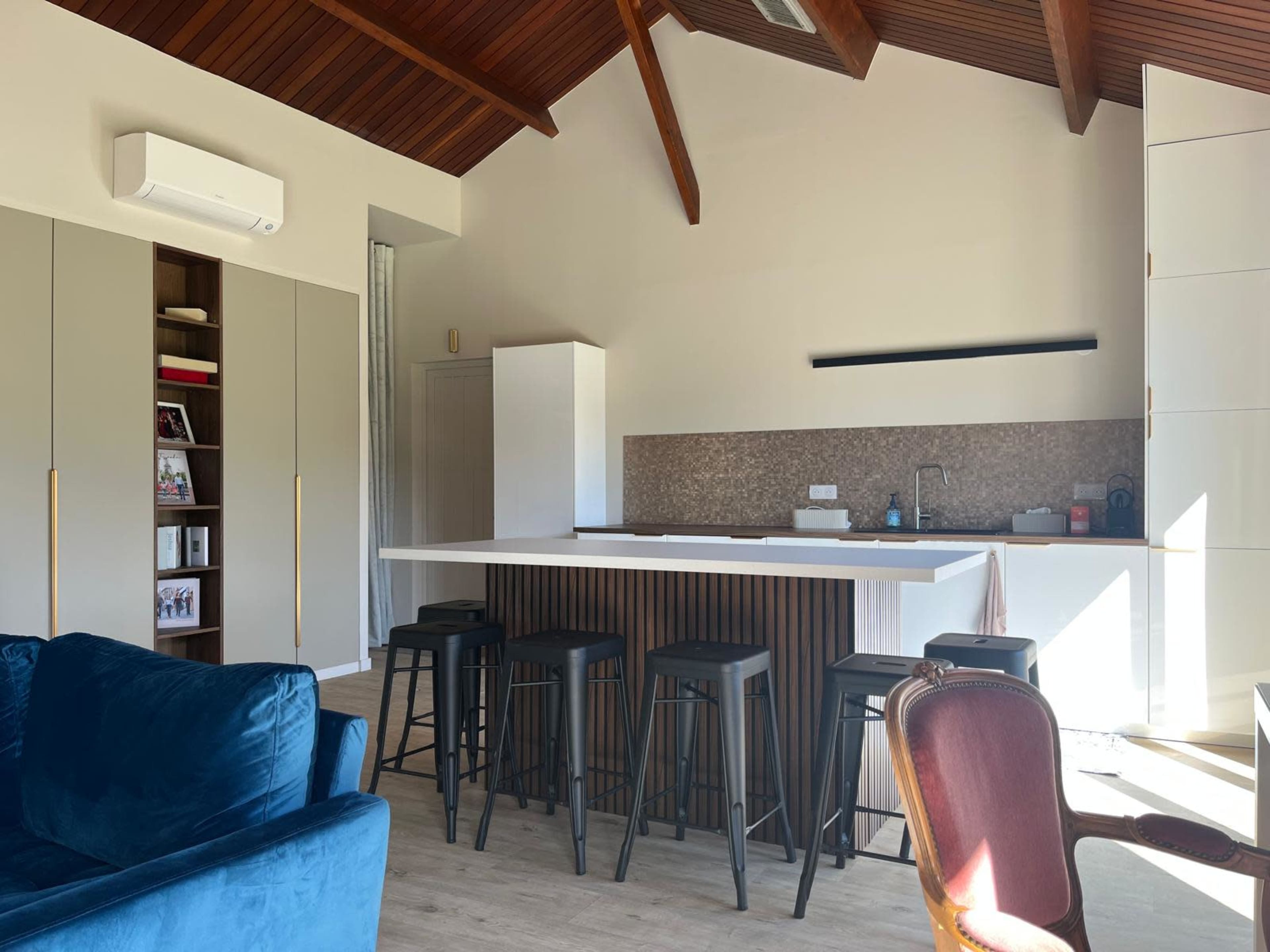 The image depicts a modern kitchen with a central island and tall stools, surrounded by wooden beams and light-colored cabinetry.