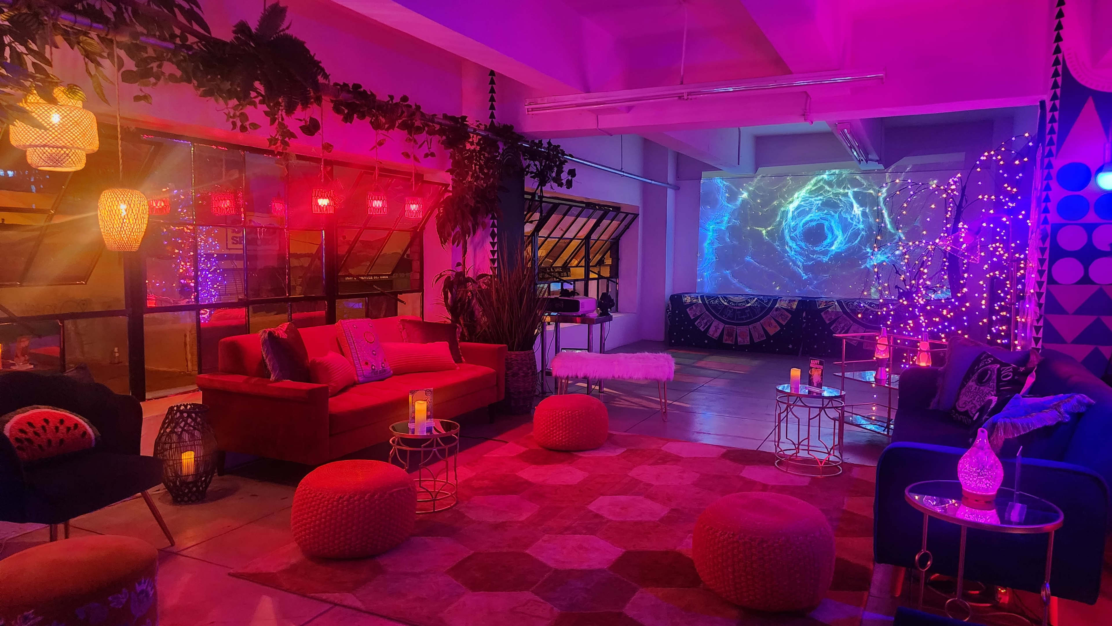 The image shows a vibrant lounge space with colorful lighting, a projector screen displaying swirling visuals, and various seating options including sofas and poufs.
