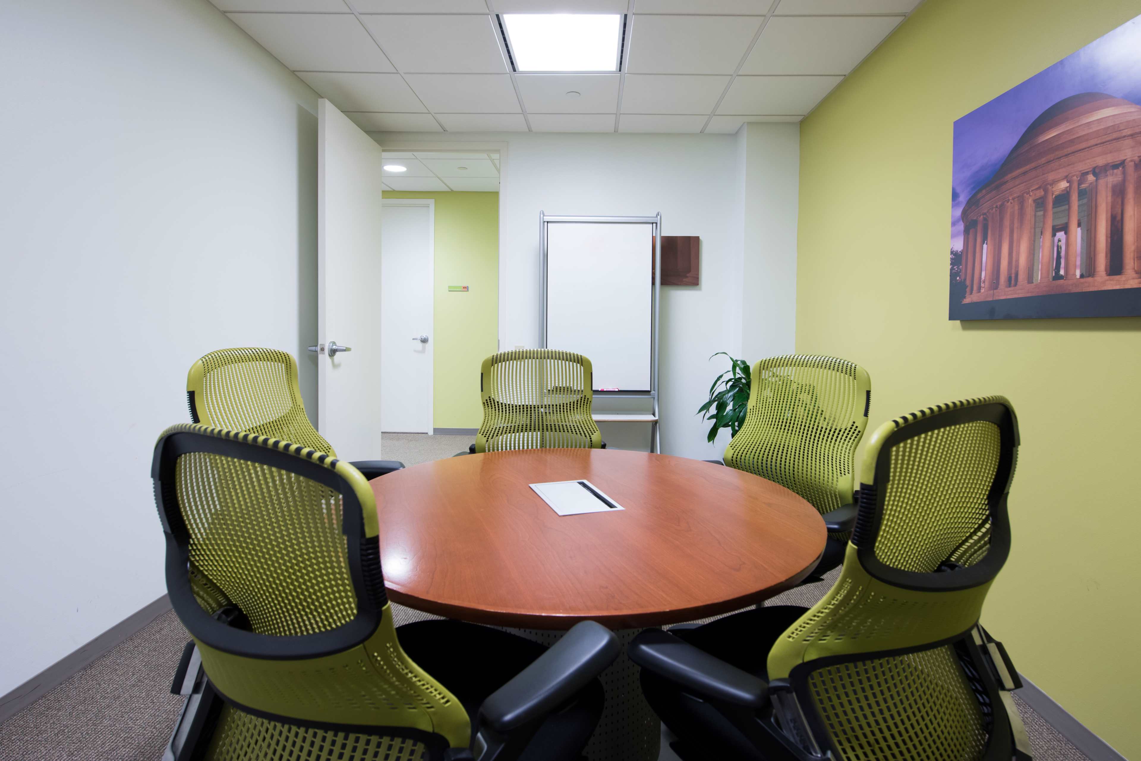 A small conference room features a round wooden table surrounded by four green mesh chairs, with a whiteboard and a framed picture on the wall.