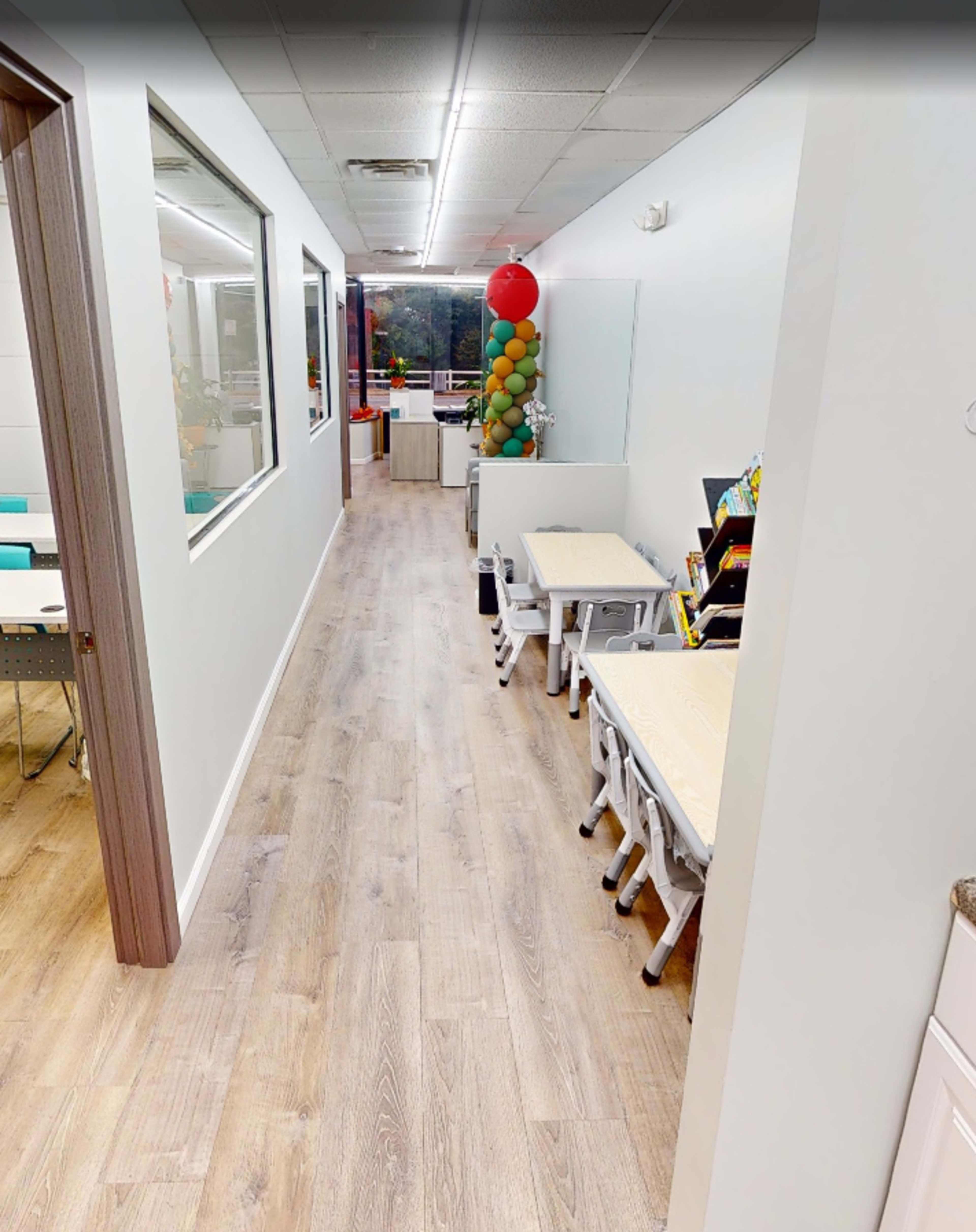 The image shows a bright hallway with white walls, wooden flooring, and a row of desks on one side, leading to a reception area decorated with colorful balloons.