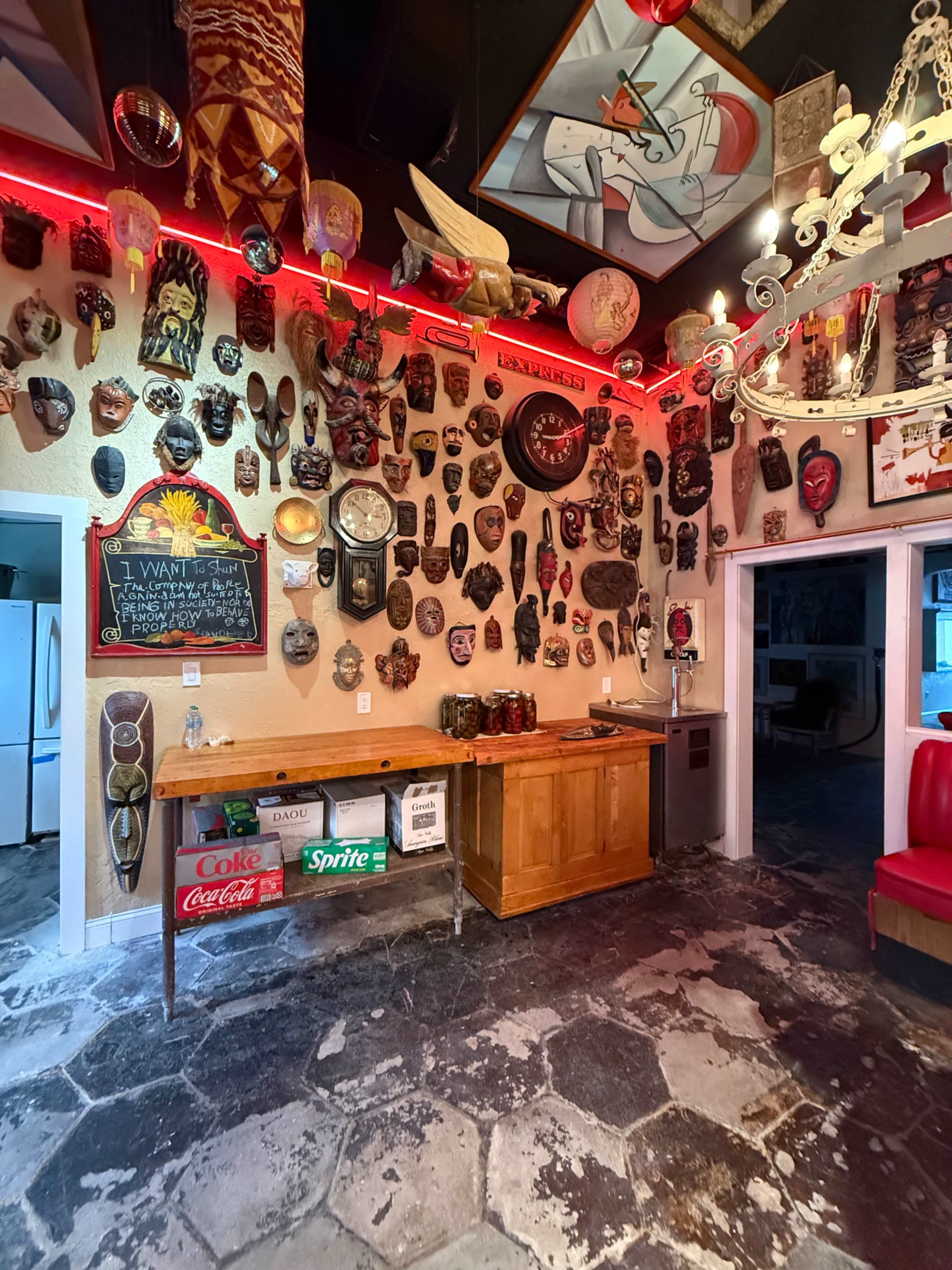 The image features a room with a wooden bar against a wall adorned with various masks and artistic decorations, illuminated by red lighting.