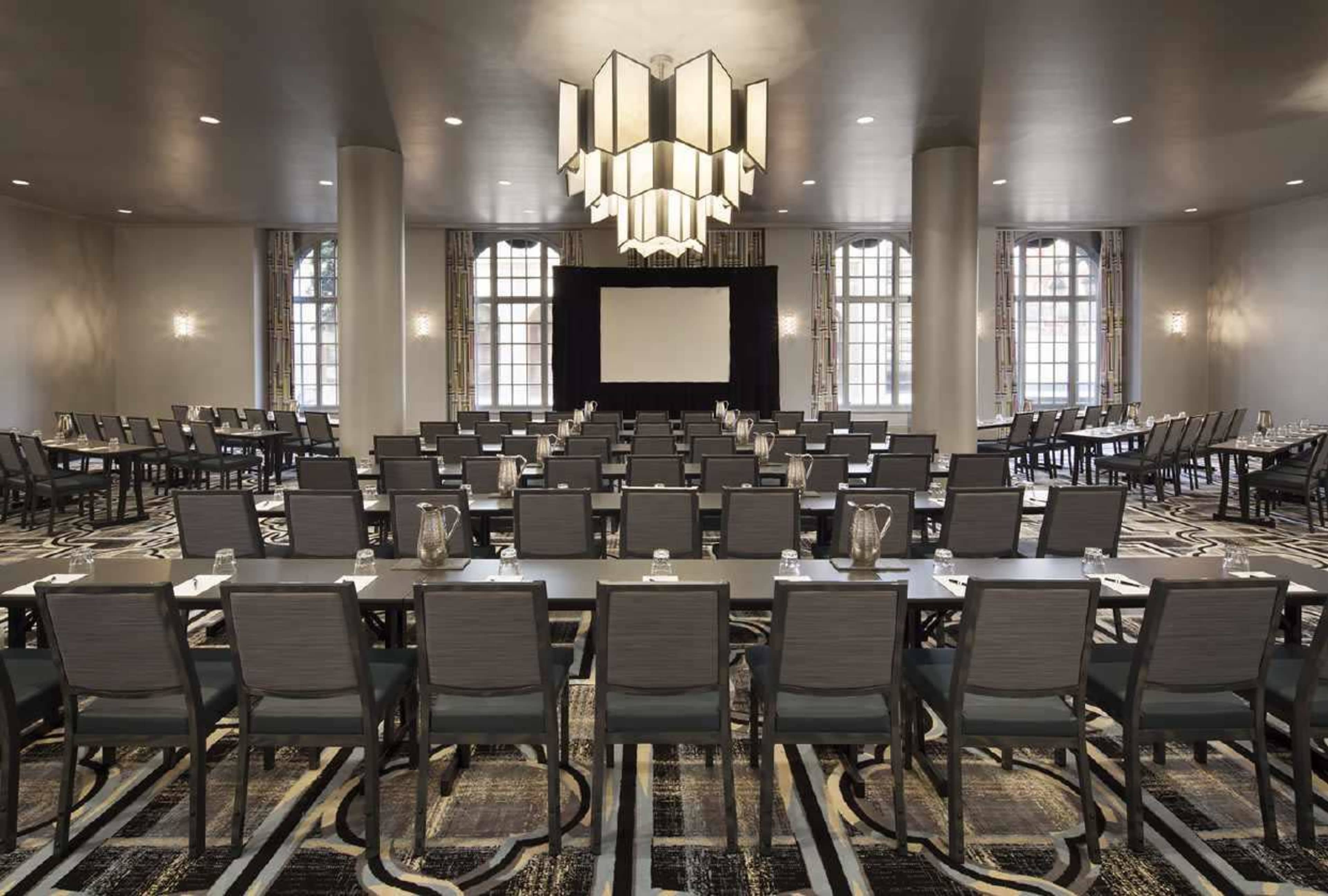 A conference room is arranged with rows of tables and chairs facing a large screen, illuminated by a modern chandelier.