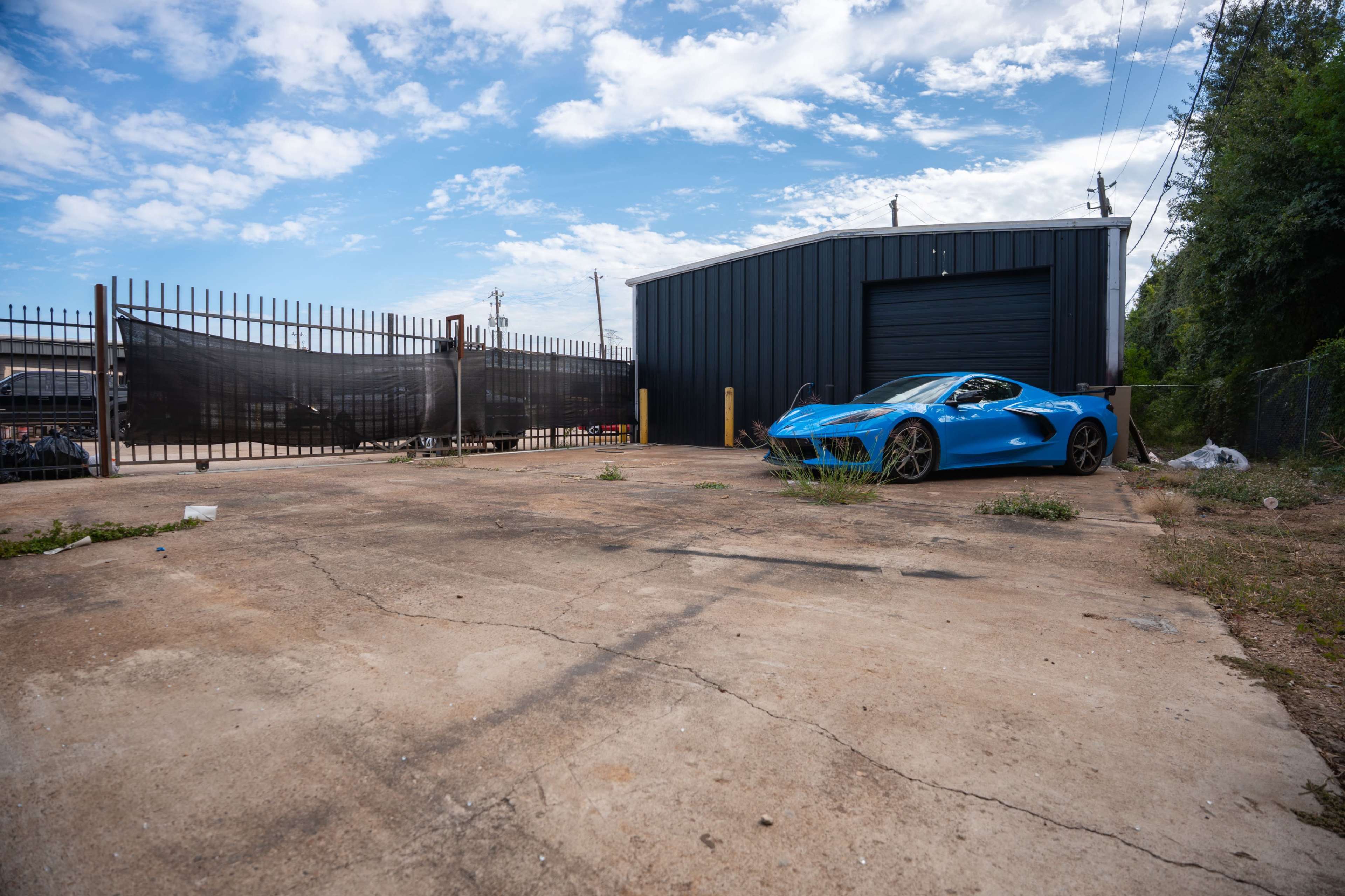 A blue sports car is parked on an empty concrete lot next to a black metal building under a partly cloudy sky.