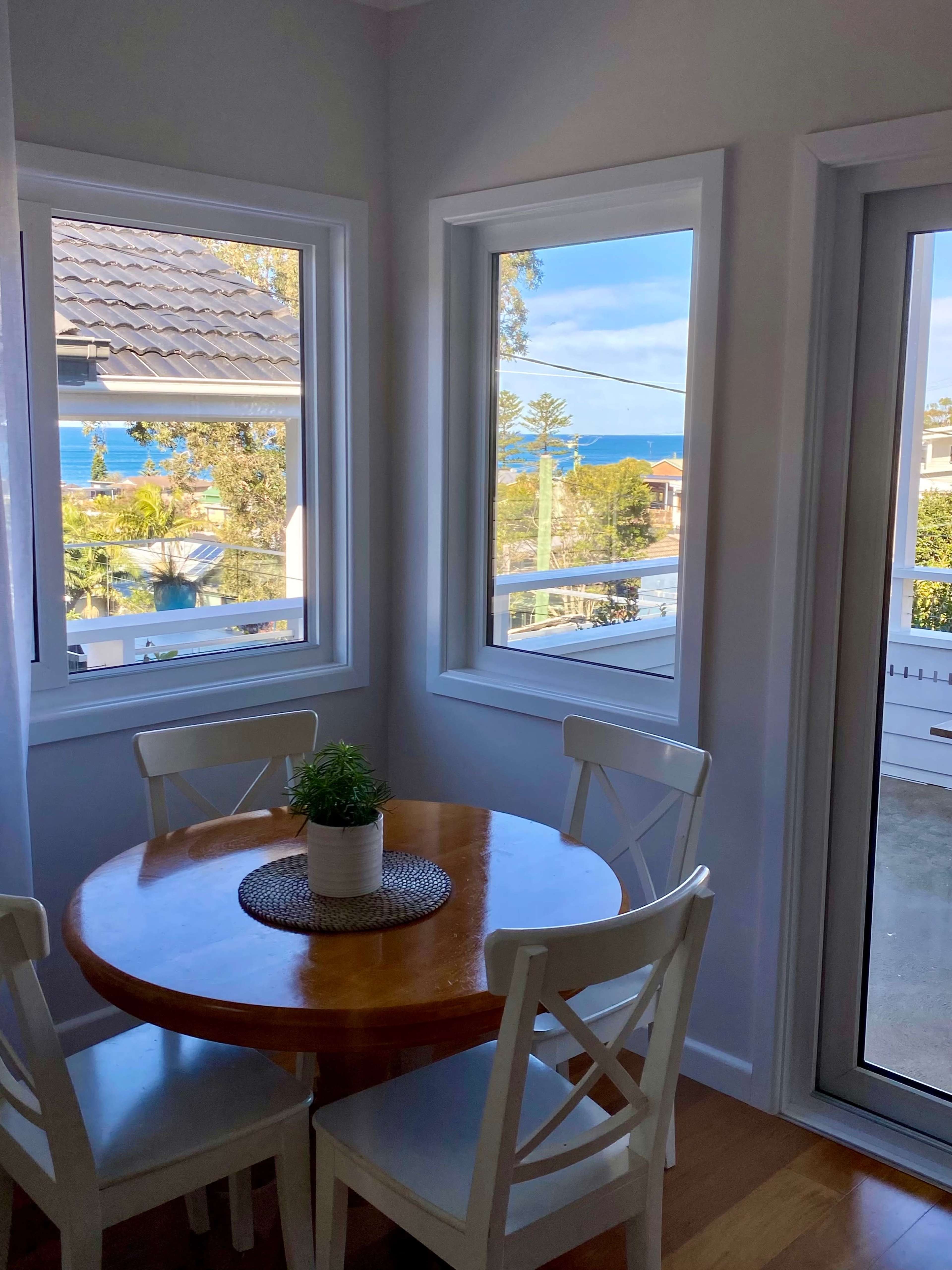 A round wooden table with four white chairs is set against a backdrop of large windows overlooking a coastal view.