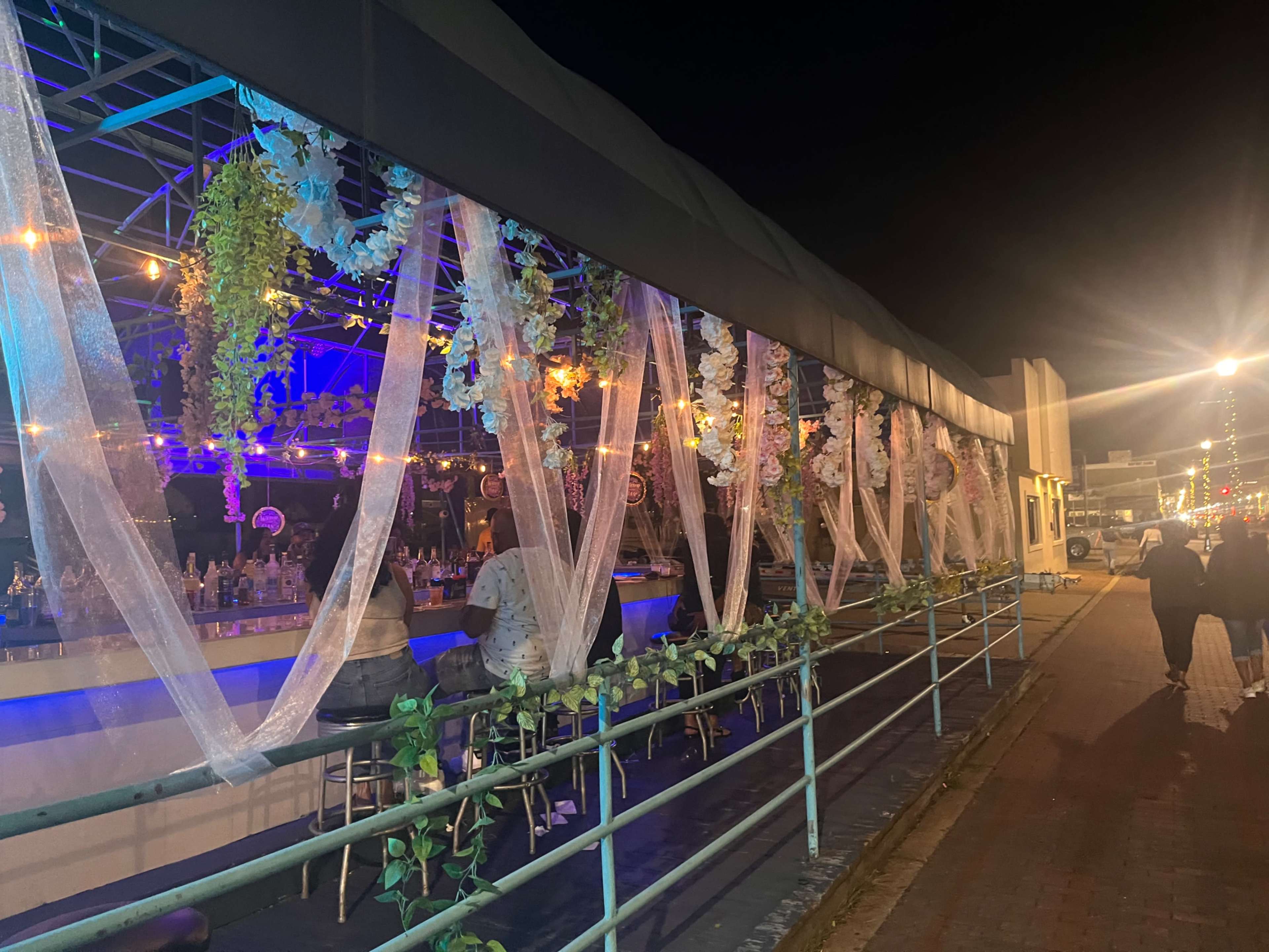 The image shows a bar with a decorated exterior lit by colorful lights at night, with people sitting at the bar and walking along the sidewalk.