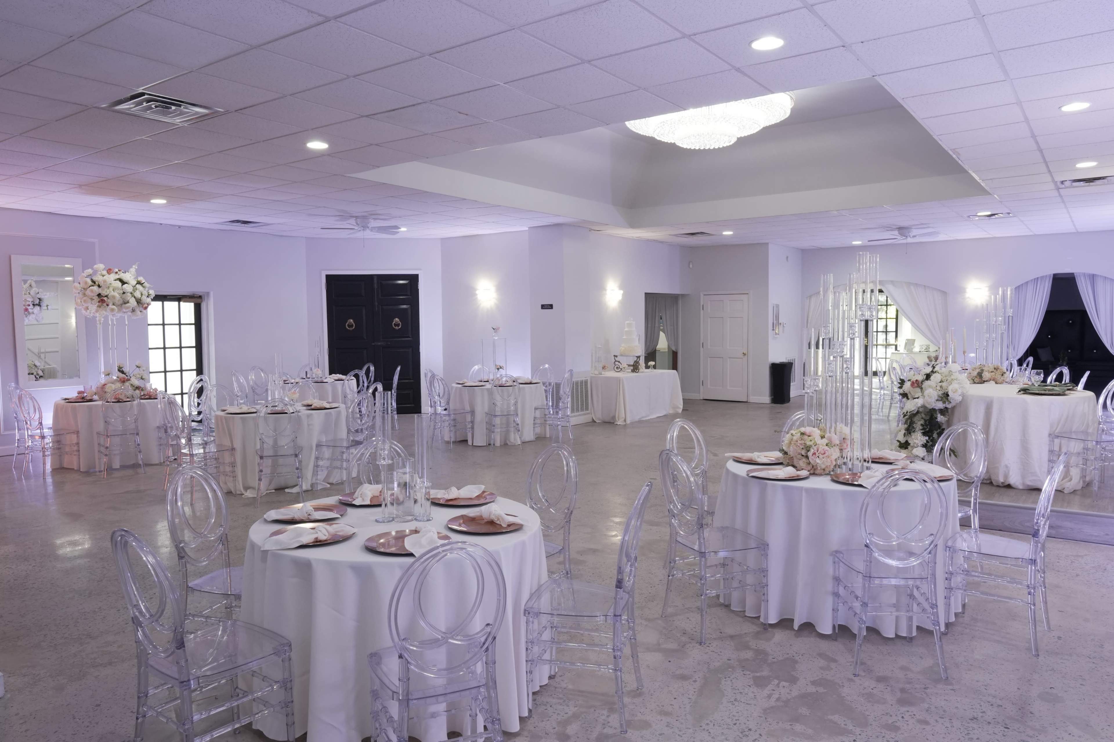 The image shows an elegantly decorated banquet hall with round tables set for an event, featuring clear acrylic chairs and floral centerpieces.