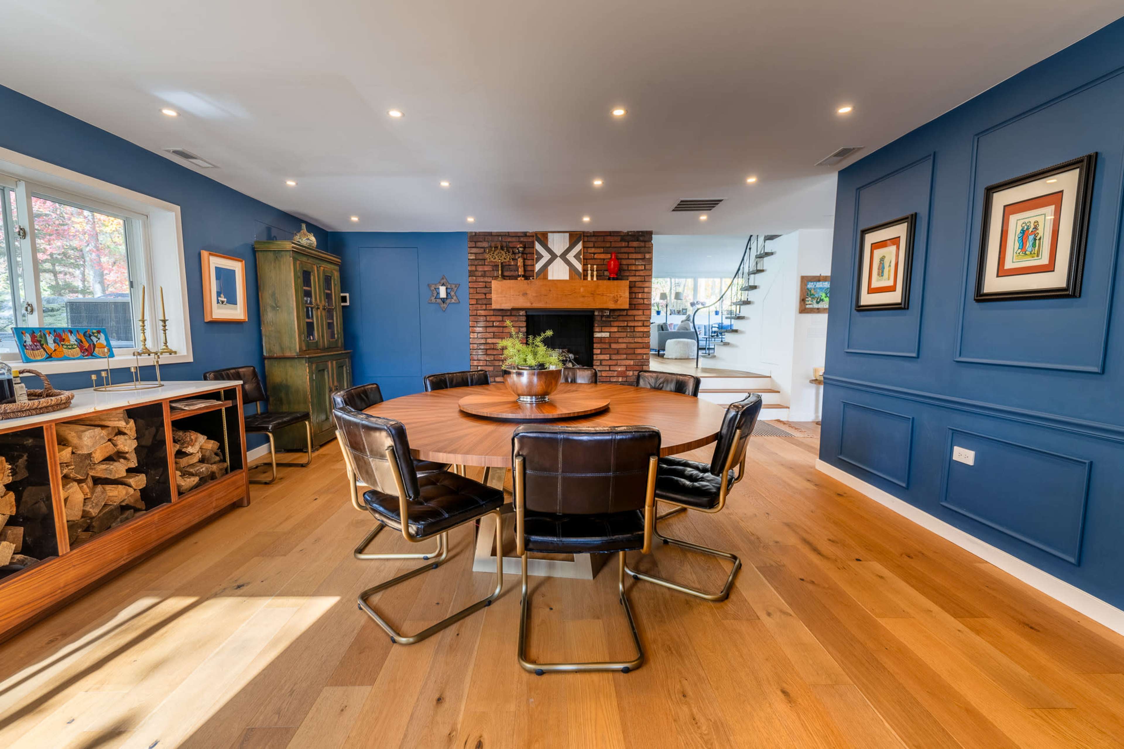 A circular wooden dining table surrounded by black metal chairs is positioned in a room with blue walls and a fireplace, illuminated by recessed lighting.