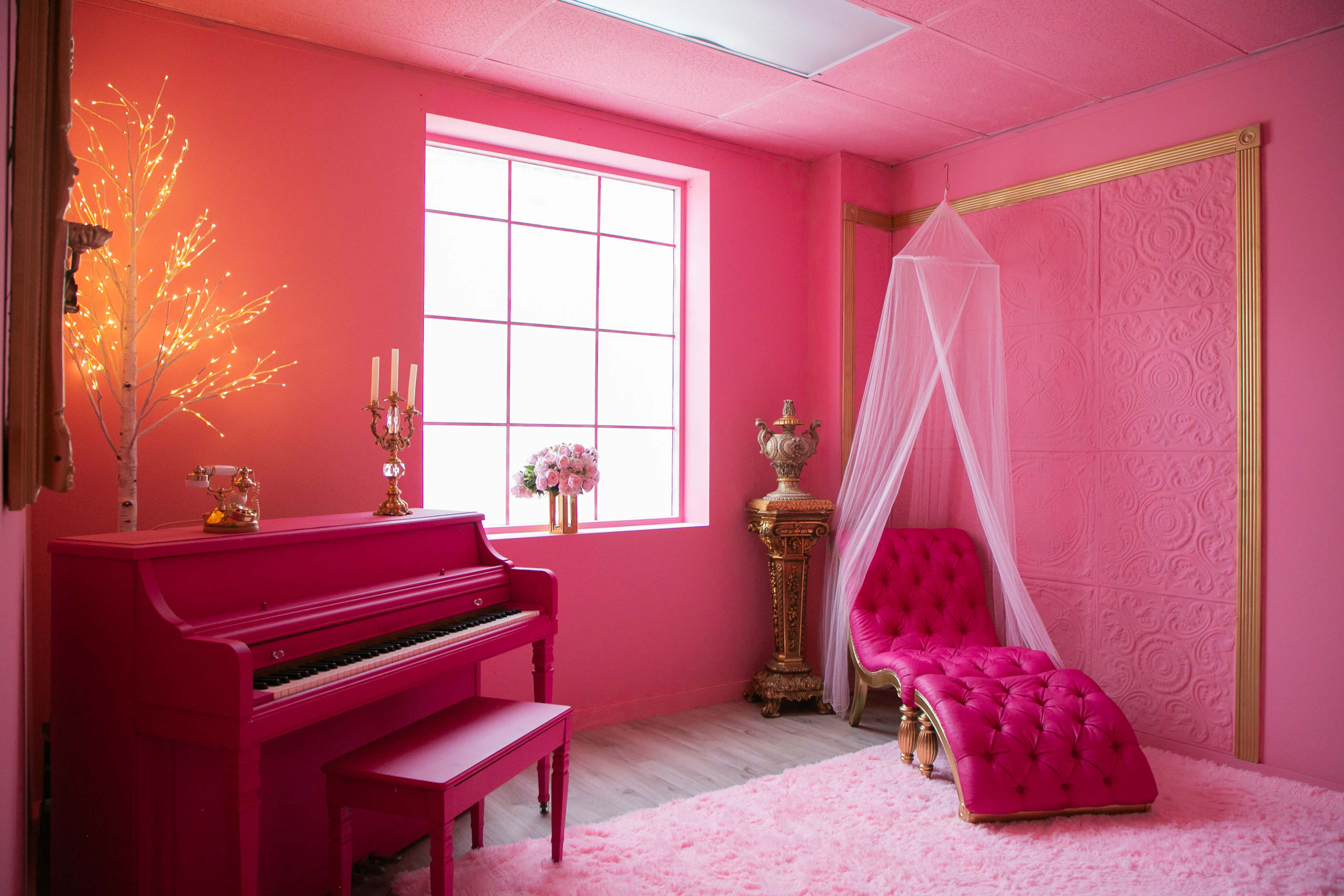The room features a bright pink color scheme with a pink piano, a tufted chaise lounge, and decorative elements like fairy lights and a floral arrangement.