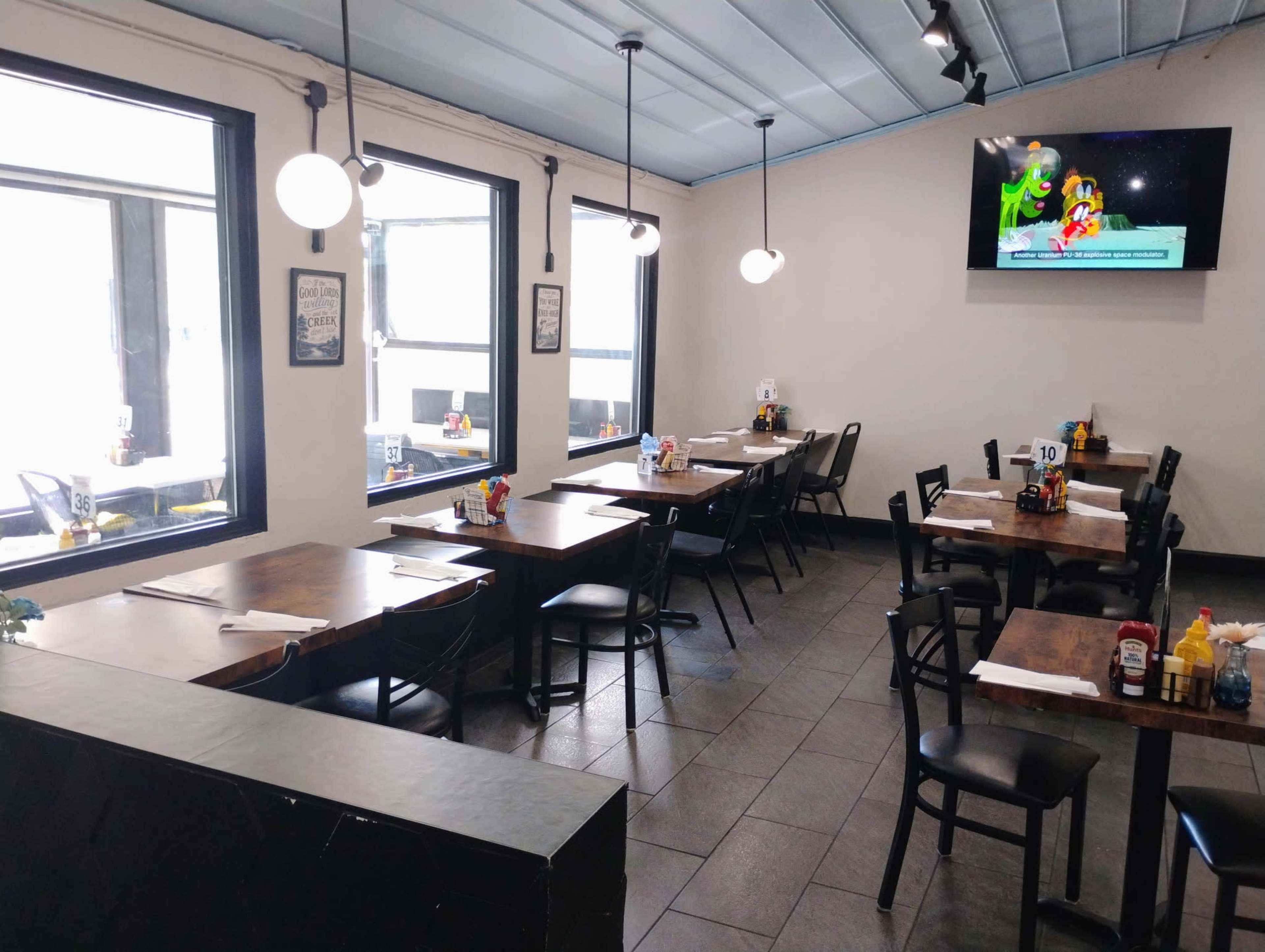 The image shows a spacious restaurant interior with several tables set for dining, black chairs, and a television displaying animated characters on the wall.