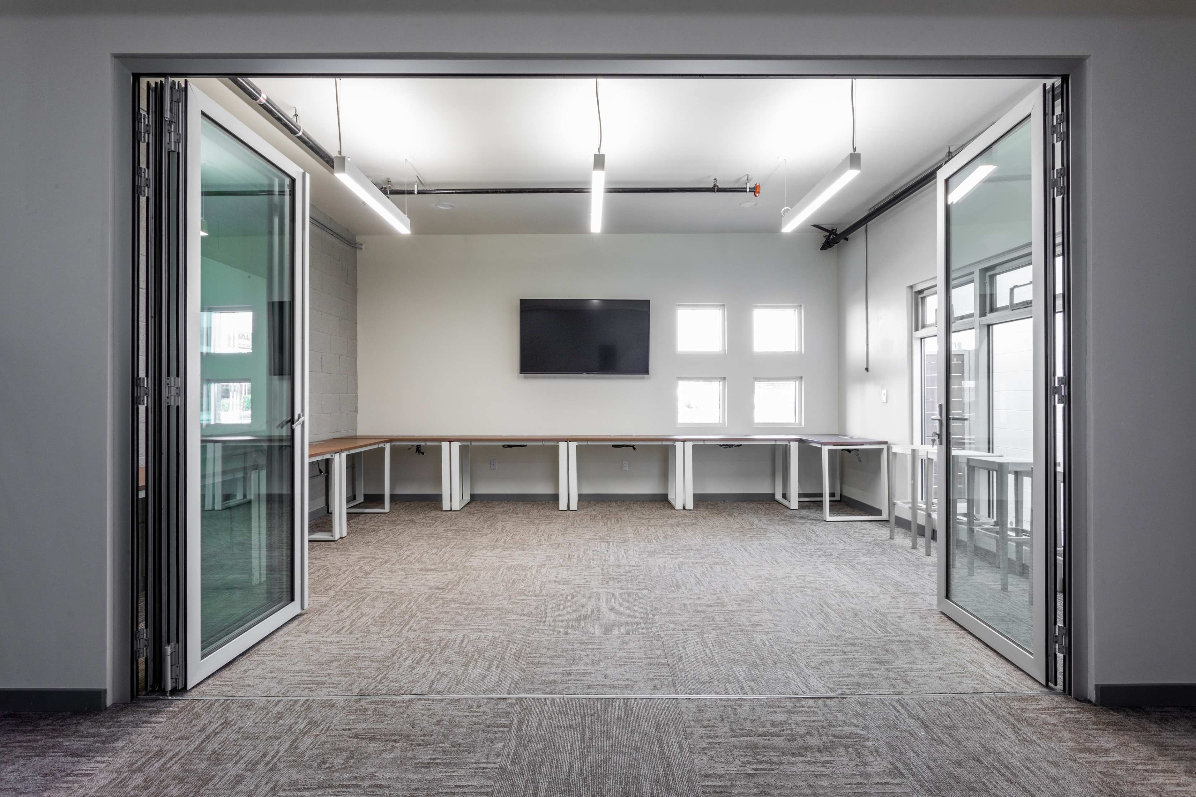 A spacious meeting room features glass doors, a large table, overhead lights, and a wall-mounted screen.