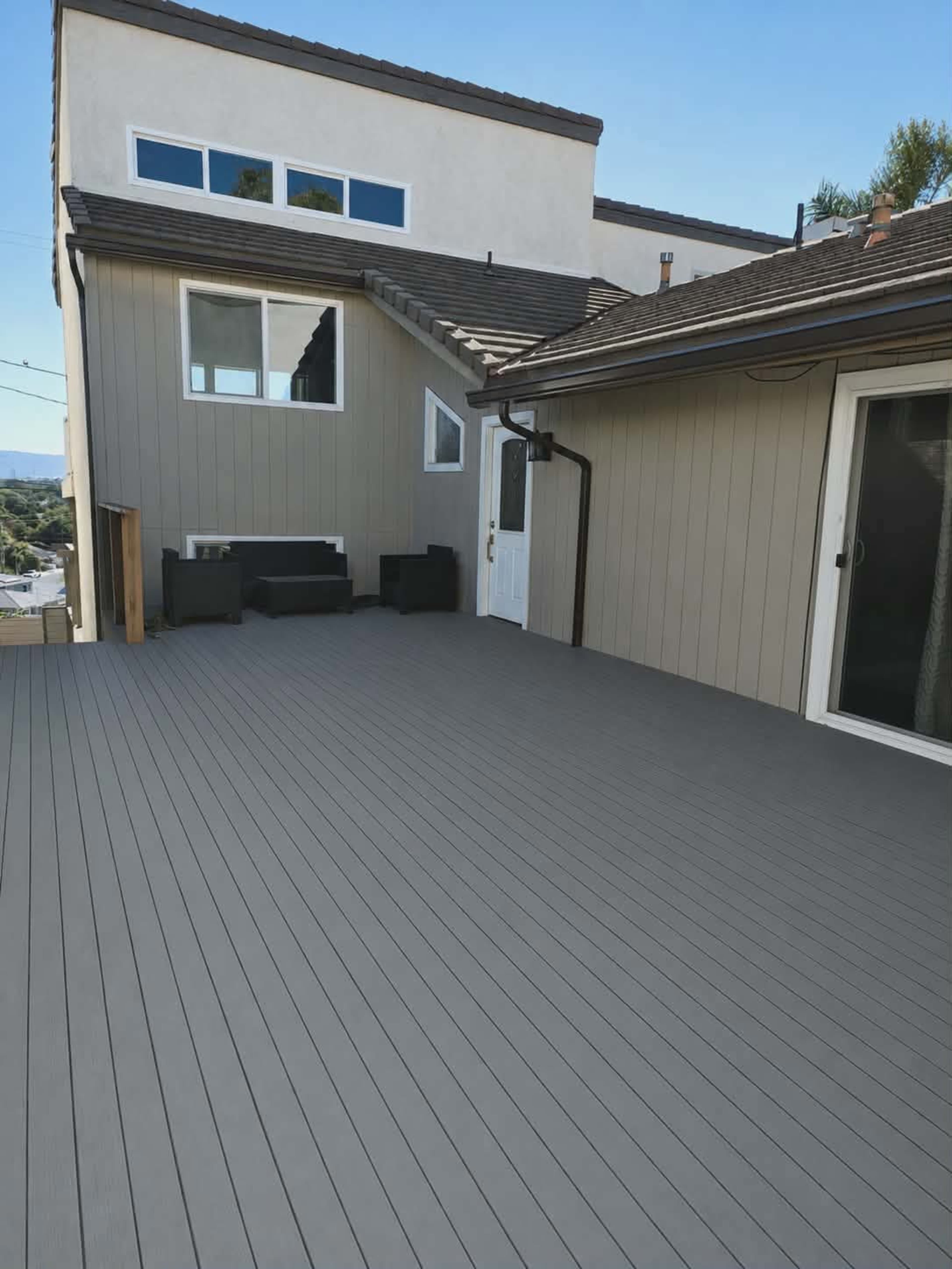 The image shows a spacious outdoor deck with a grey composite floor, adjacent to a house featuring a light-colored exterior, a door, and a seating area.