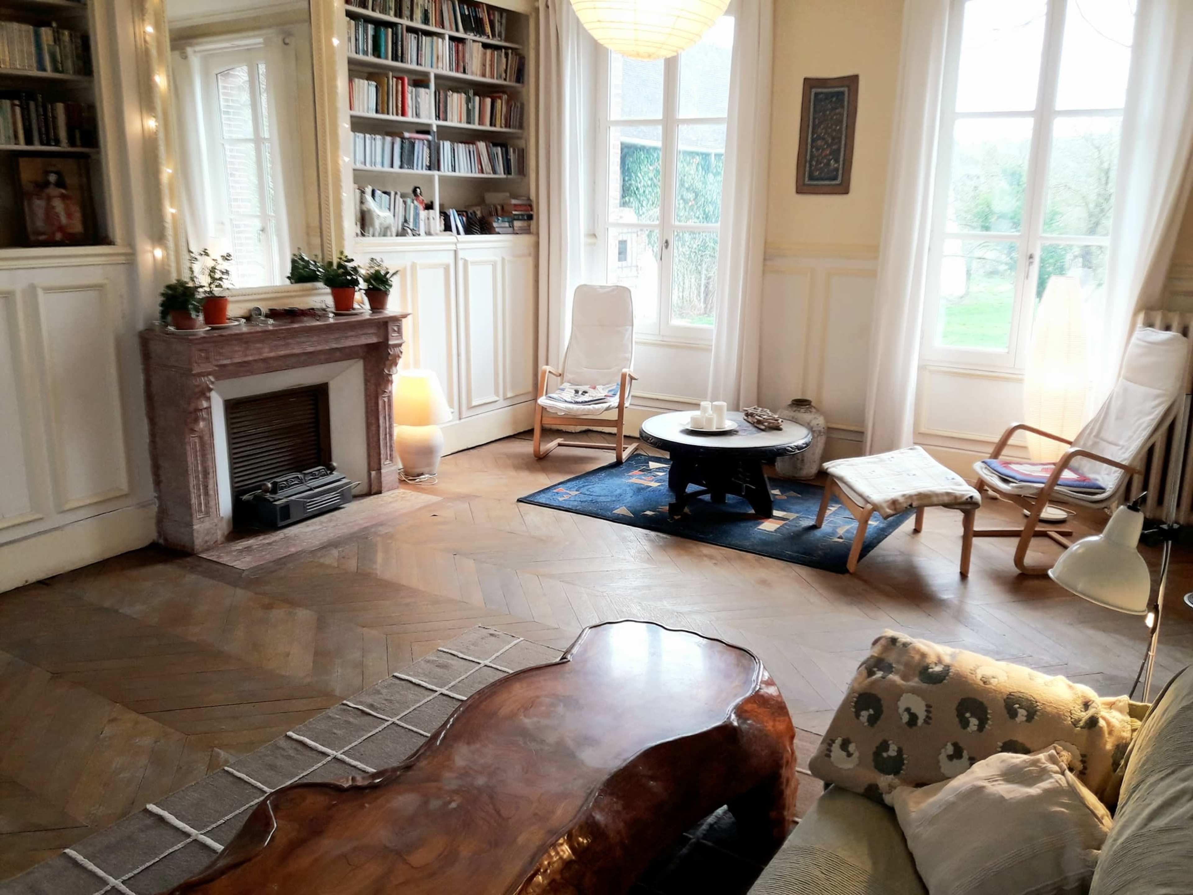 A cozy living room with hardwood floors, bookshelves, armchairs, and large windows allowing natural light.