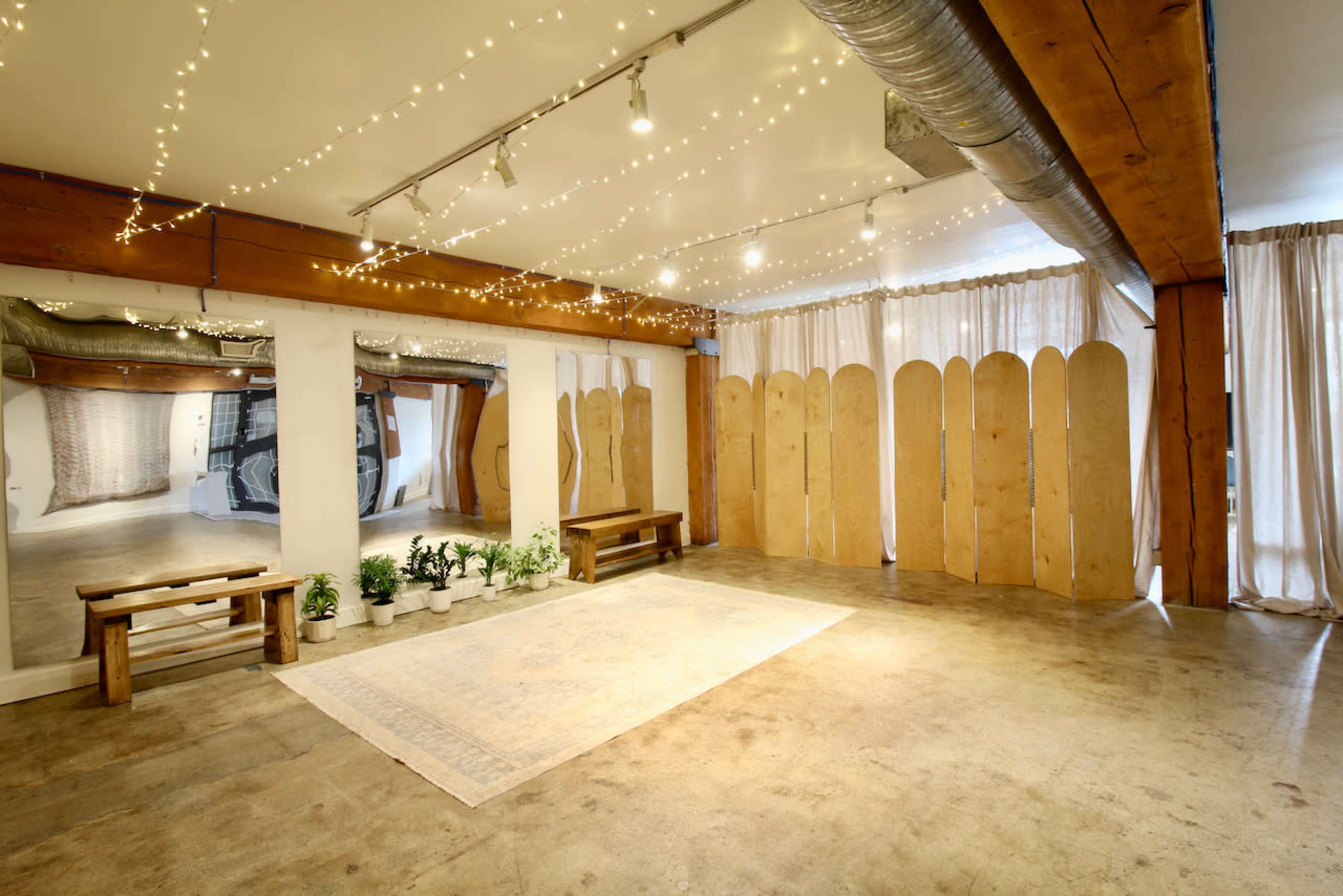 The image shows a spacious, minimally decorated indoor area with wooden beams, benches, a patterned rug, and soft ambient lighting from string lights.