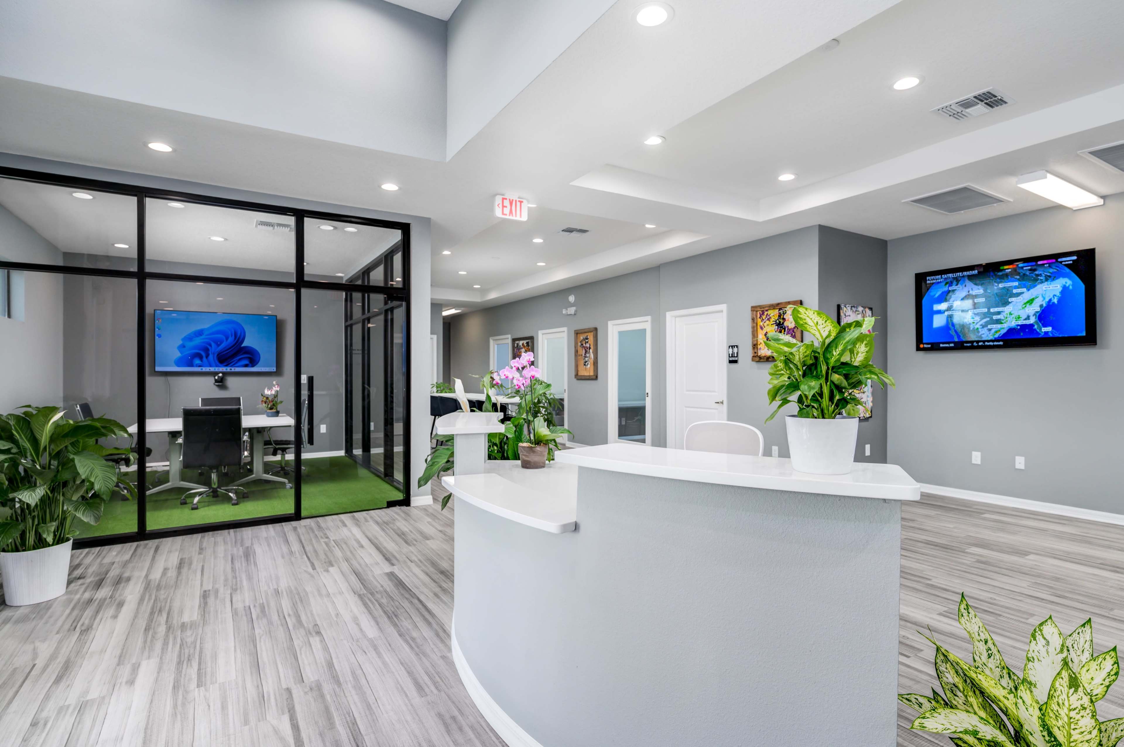 A modern office reception area with a front desk, plant decorations, and a glass-walled meeting room in the background.