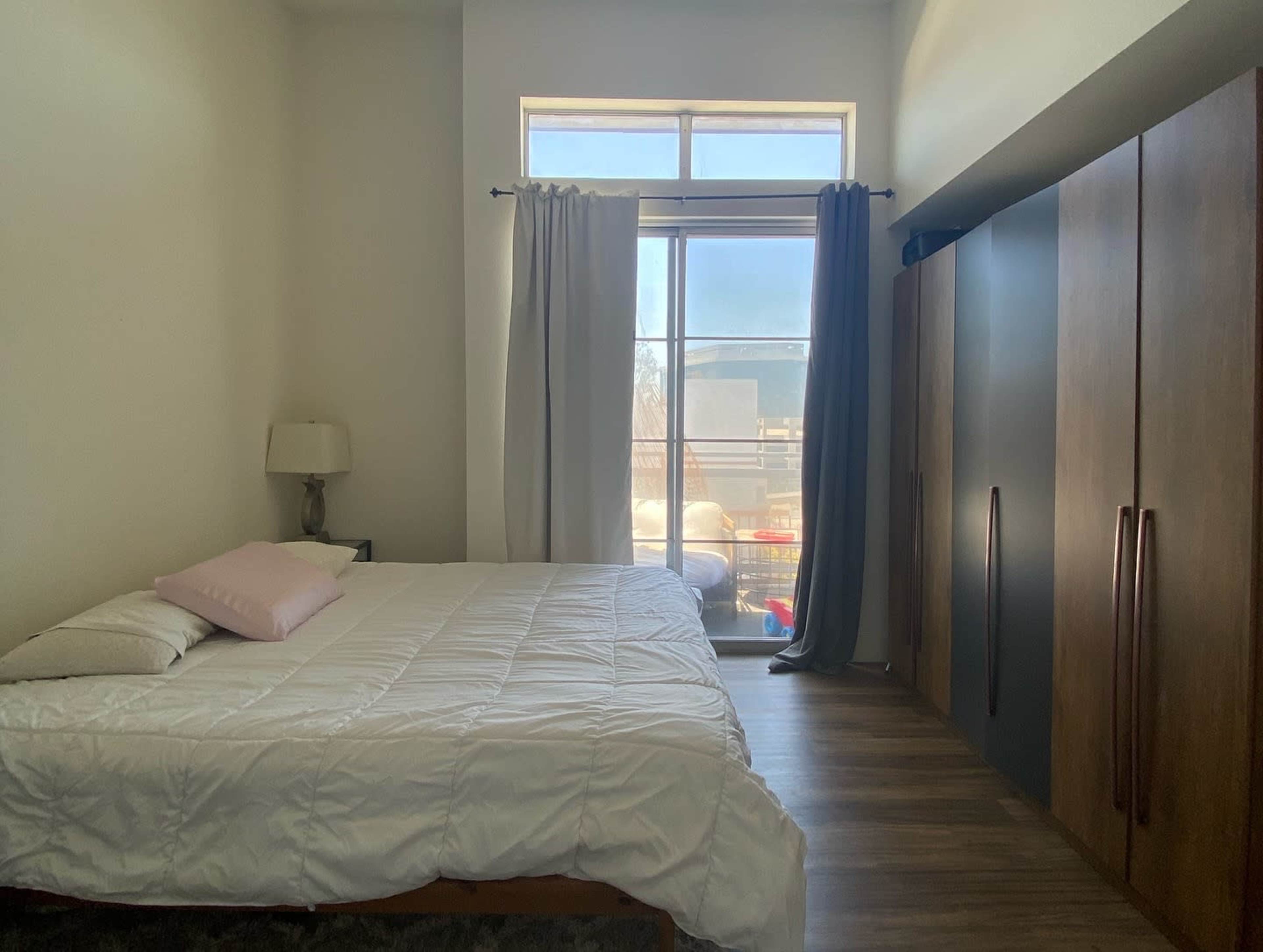 A bedroom features a large window with sheer curtains, a bed with white bedding and a pink pillow, and a wooden wardrobe.