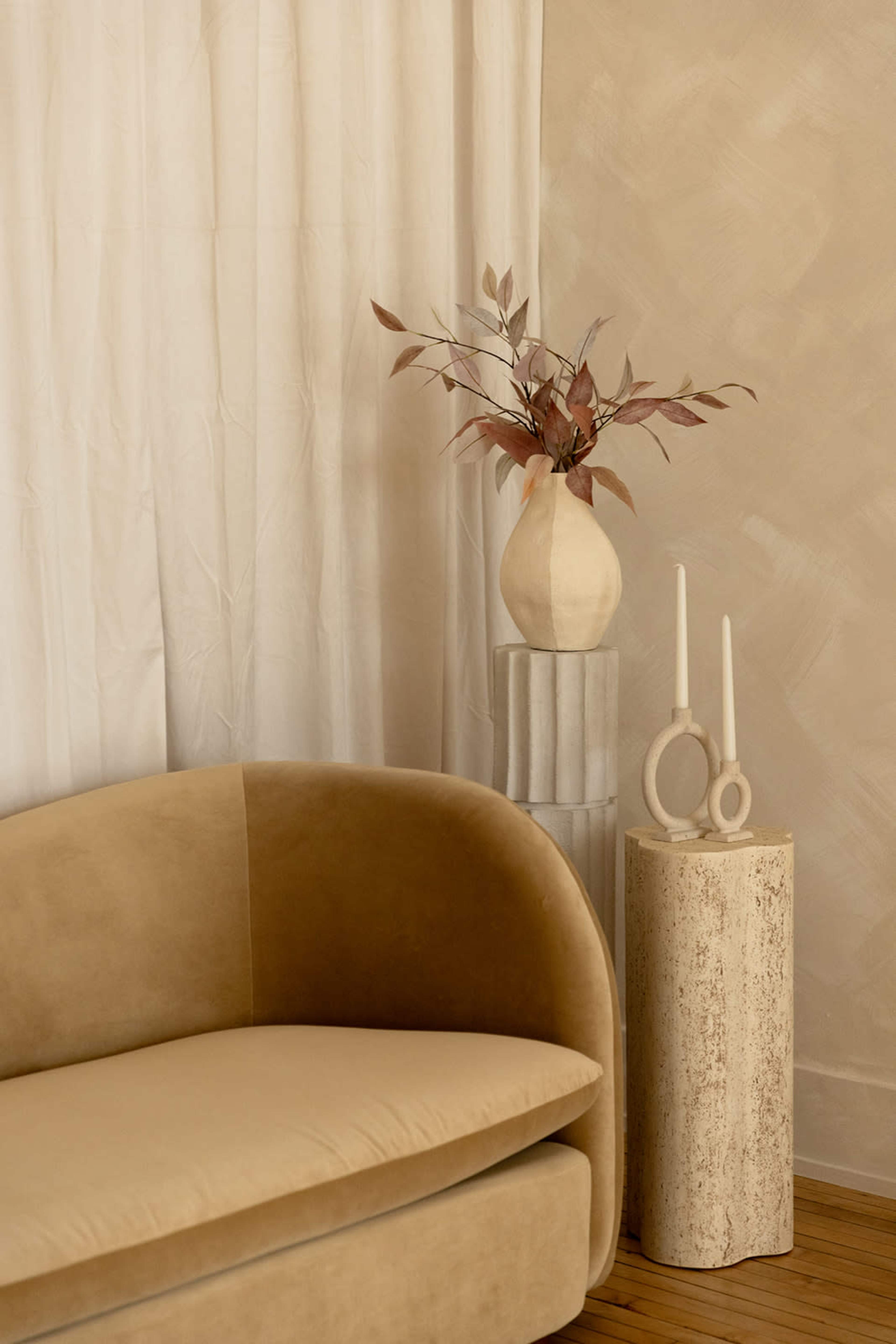 A curved beige sofa sits beside a textured stone pedestal with a decorative vase and two candlesticks, against a backdrop of white curtains and a soft beige wall.
