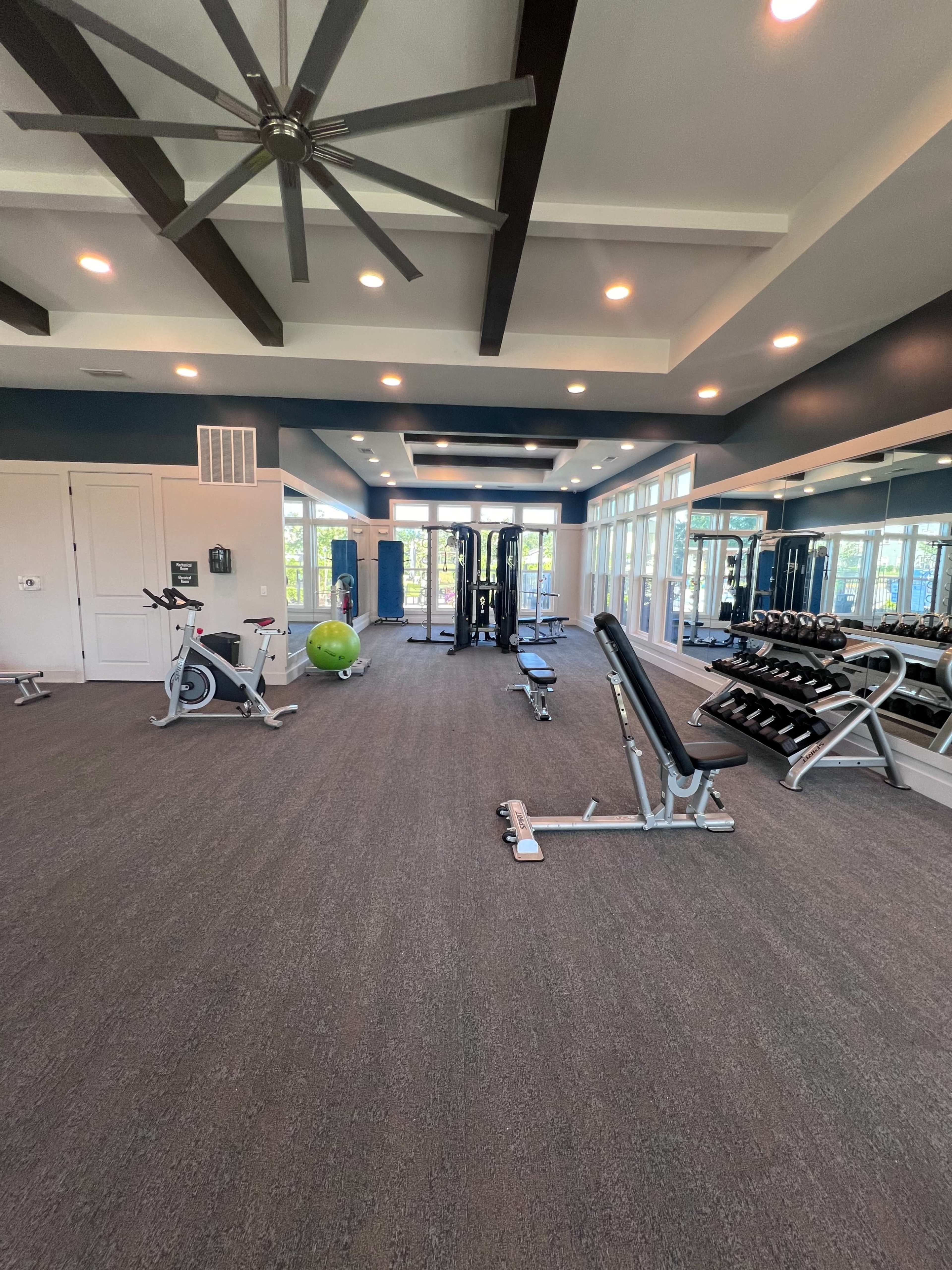 The image shows a well-lit gym equipped with various exercise machines, free weights, and a large window allowing natural light to enter.